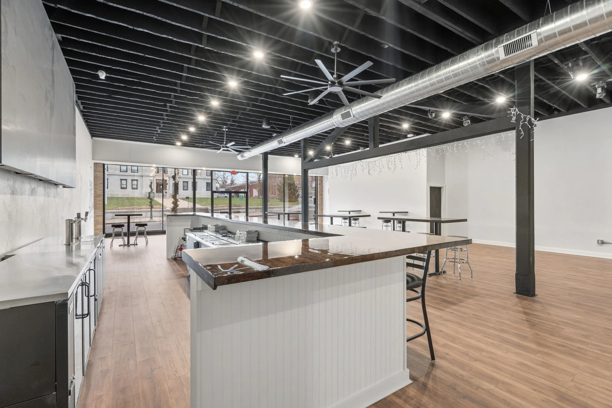 Interior of a modern cafe or restaurant with a bar counter, high stools, tables, and chairs, large front windows, black ceiling with exposed beams and ductwork, ceiling fans, and hardwood floors.