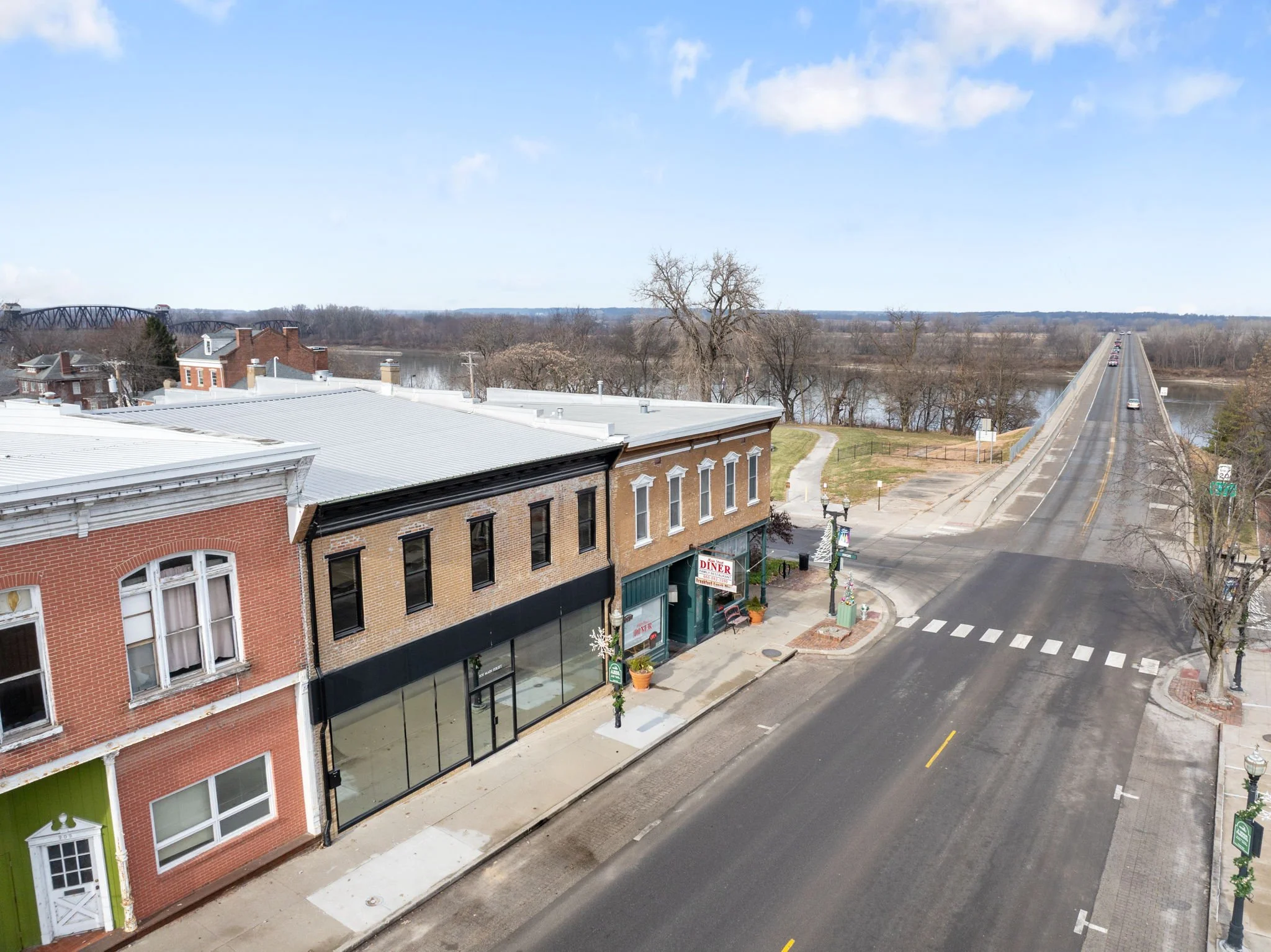 A small town street scene with historic buildings, one with large glass windows, and a restaurant sign. A bridge over a river and leafless trees are visible in the background on a clear day.