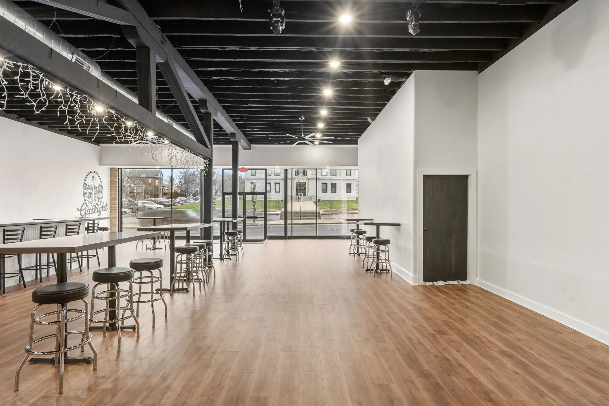 Empty coffee shop with wooden floors, high ceilings with black beams, and large windows at the front. There are bar stools along the windows and more tables and chairs on the left side. The shop has a modern, minimalist design.
