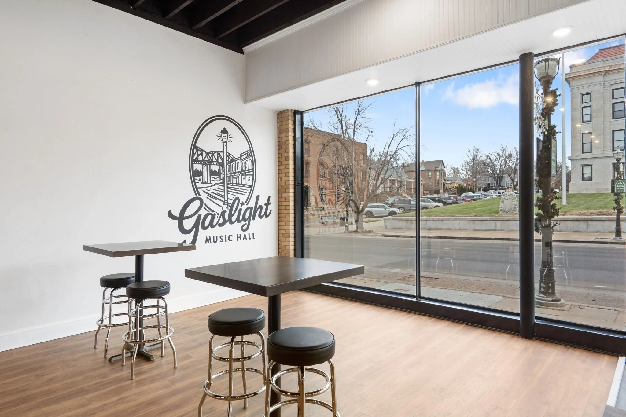 Interior of Gaslight Music Hall with high tables and bar stools near large window looking out onto a street scene with trees, parked cars, and historic buildings.