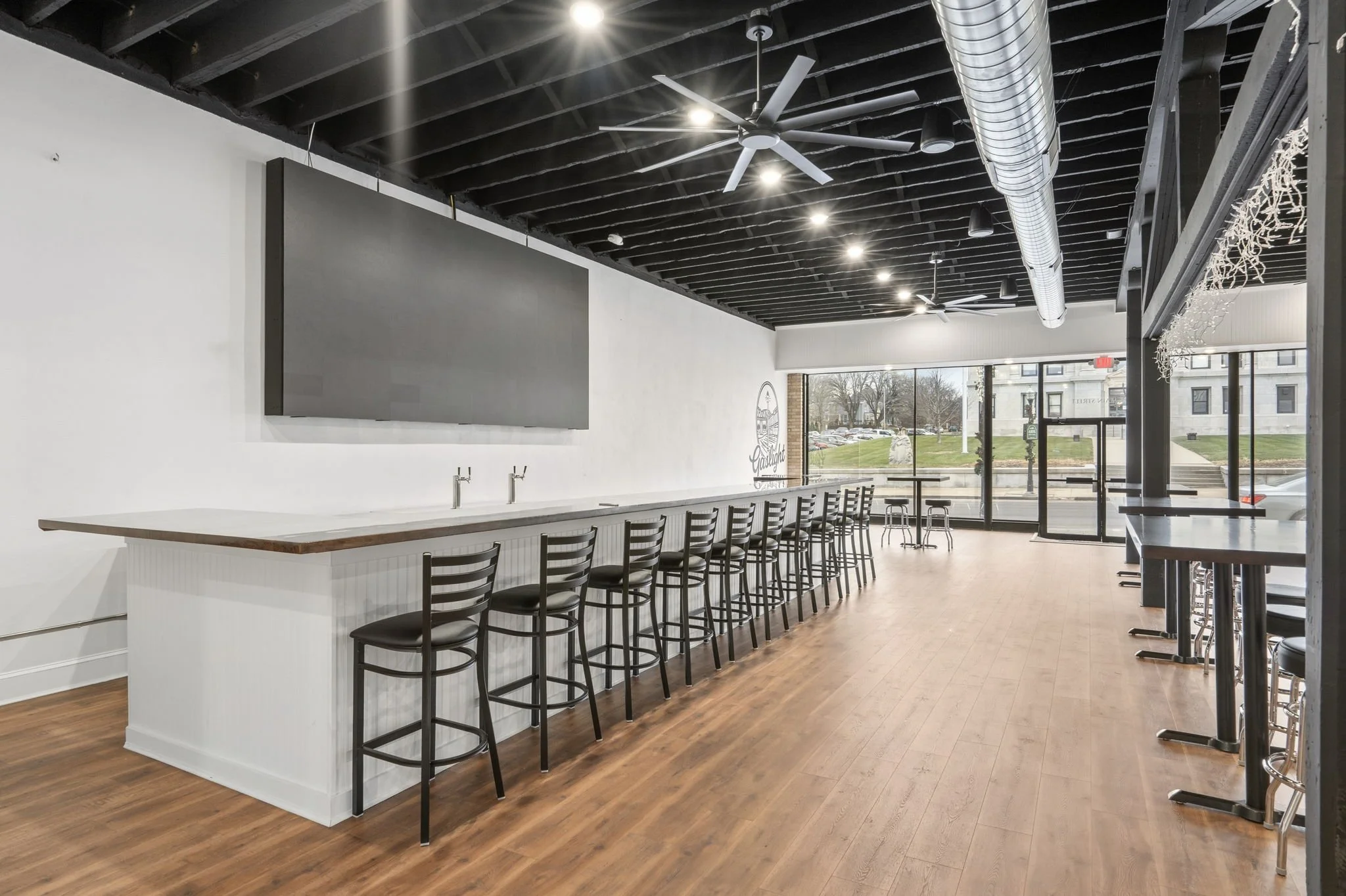 Interior of a modern casual dining or bar area with a high counter, black chairs, and large windows showing an outdoor view of a street and buildings.