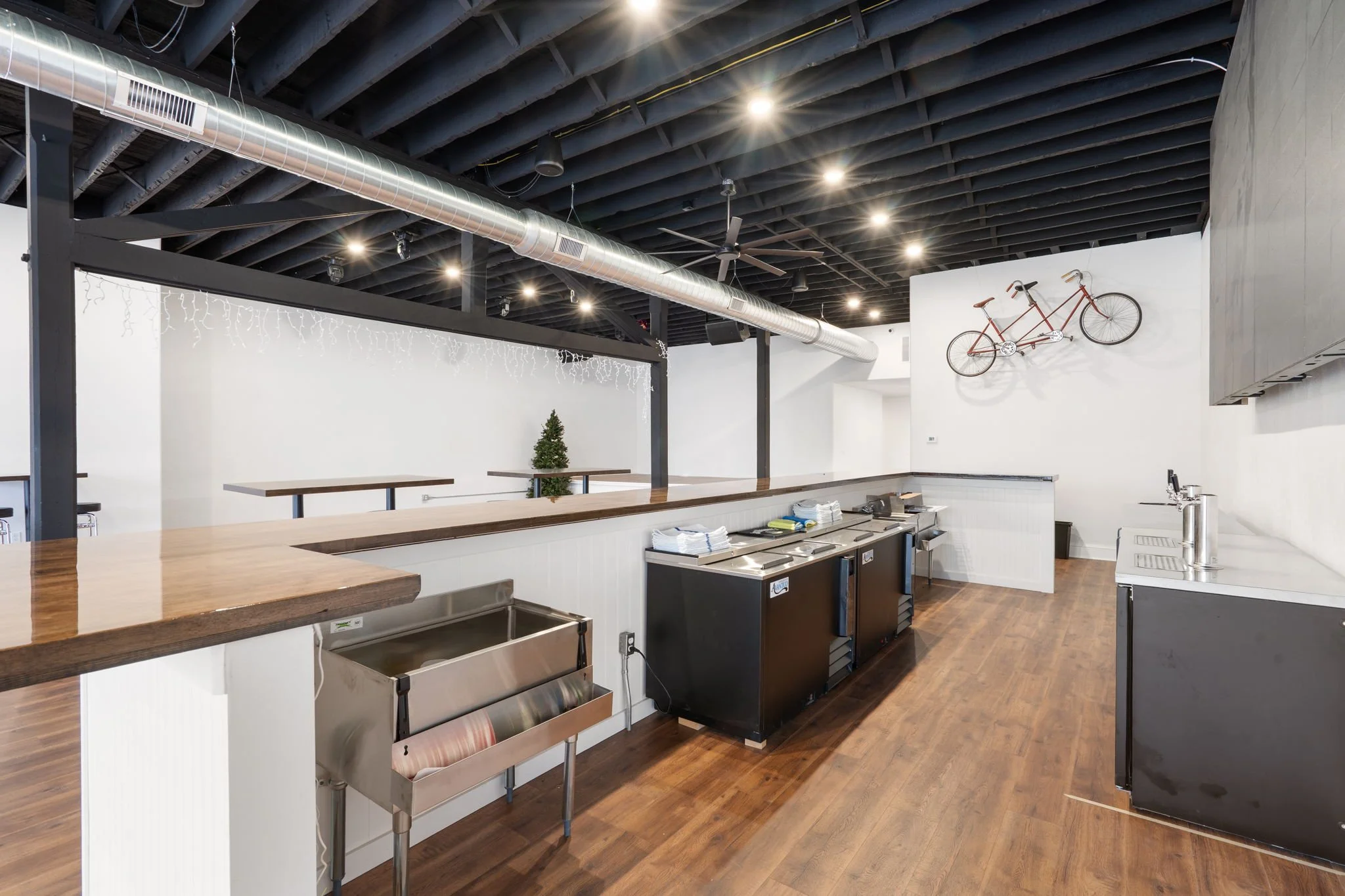 Inside view of a modern cafe or restaurant with white walls, wooden floors, and black ceiling beams featuring visible ductwork. There is a wooden counter with a metal roll-up door underneath, topped with several napkins and menus. Decorative string l