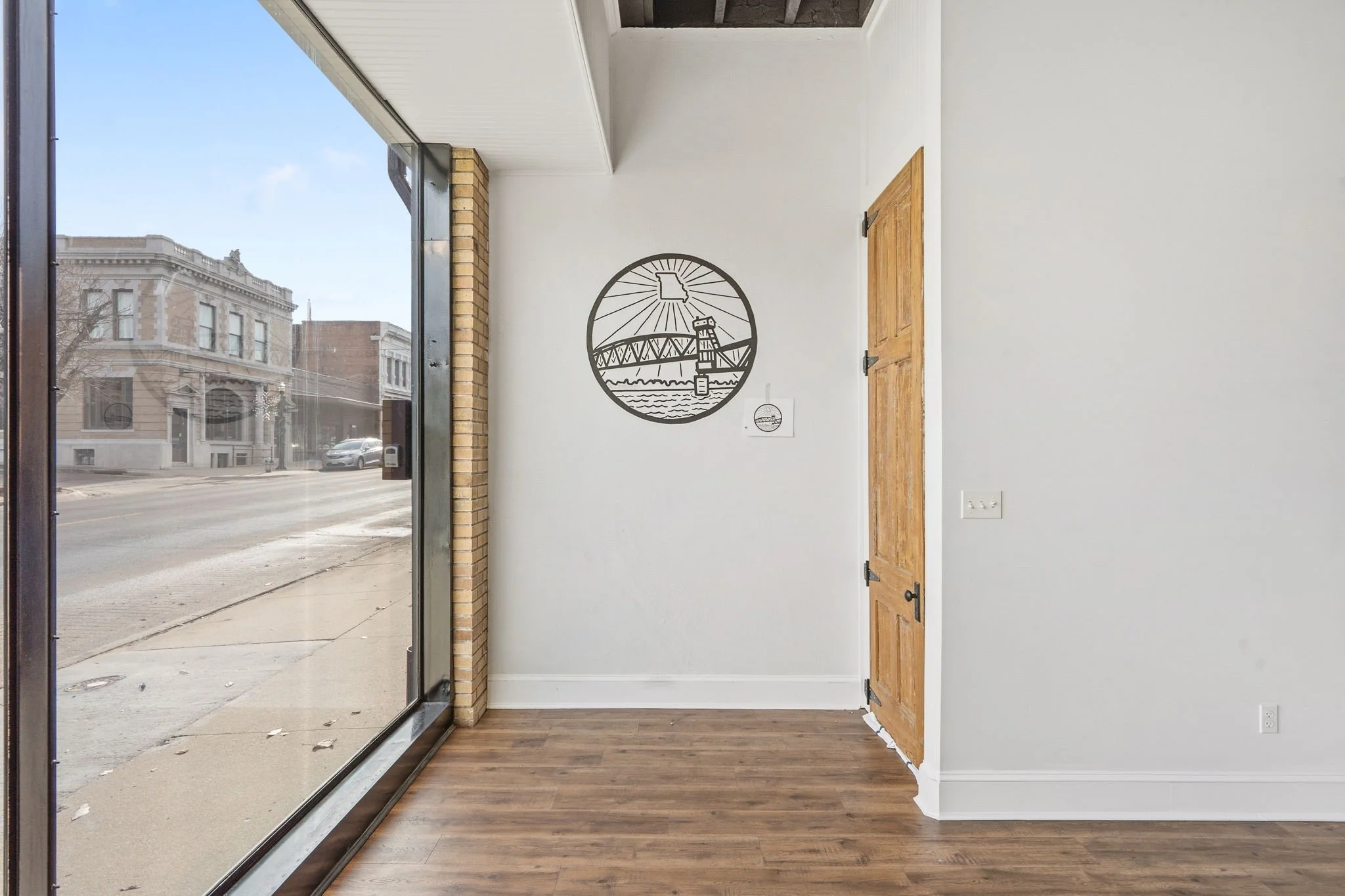 Interior of a room with hardwood floors, a large glass window showing the street outside, a white wall with a round metal wall art depicting a bridge and water, a small sign, and a wooden door.