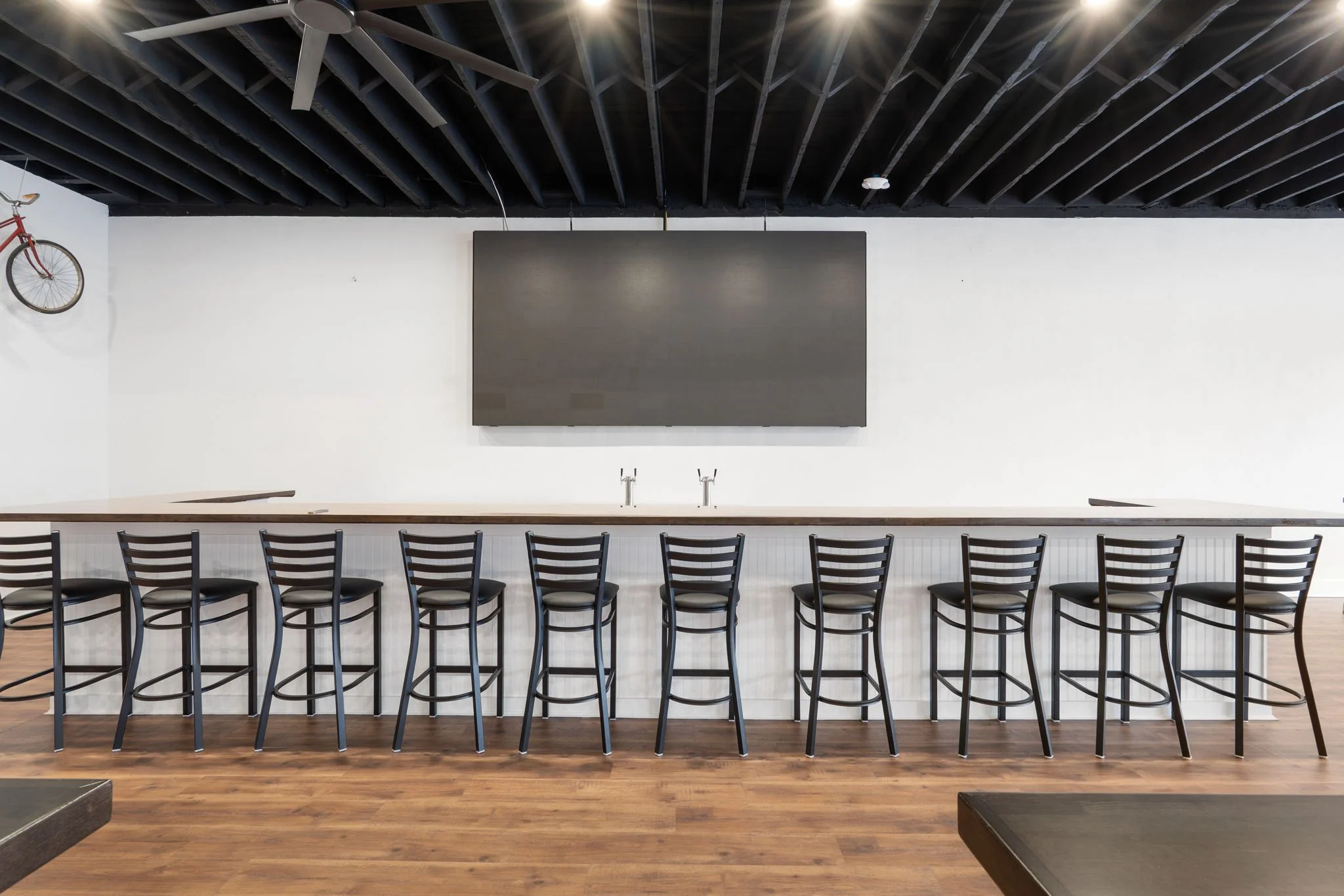 Empty bar area with nine black bar stools lining a white counter, a large blackboard on the wall, two sinks, a ceiling fan, and a red bicycle hanging on the wall in a modern space with wood flooring.