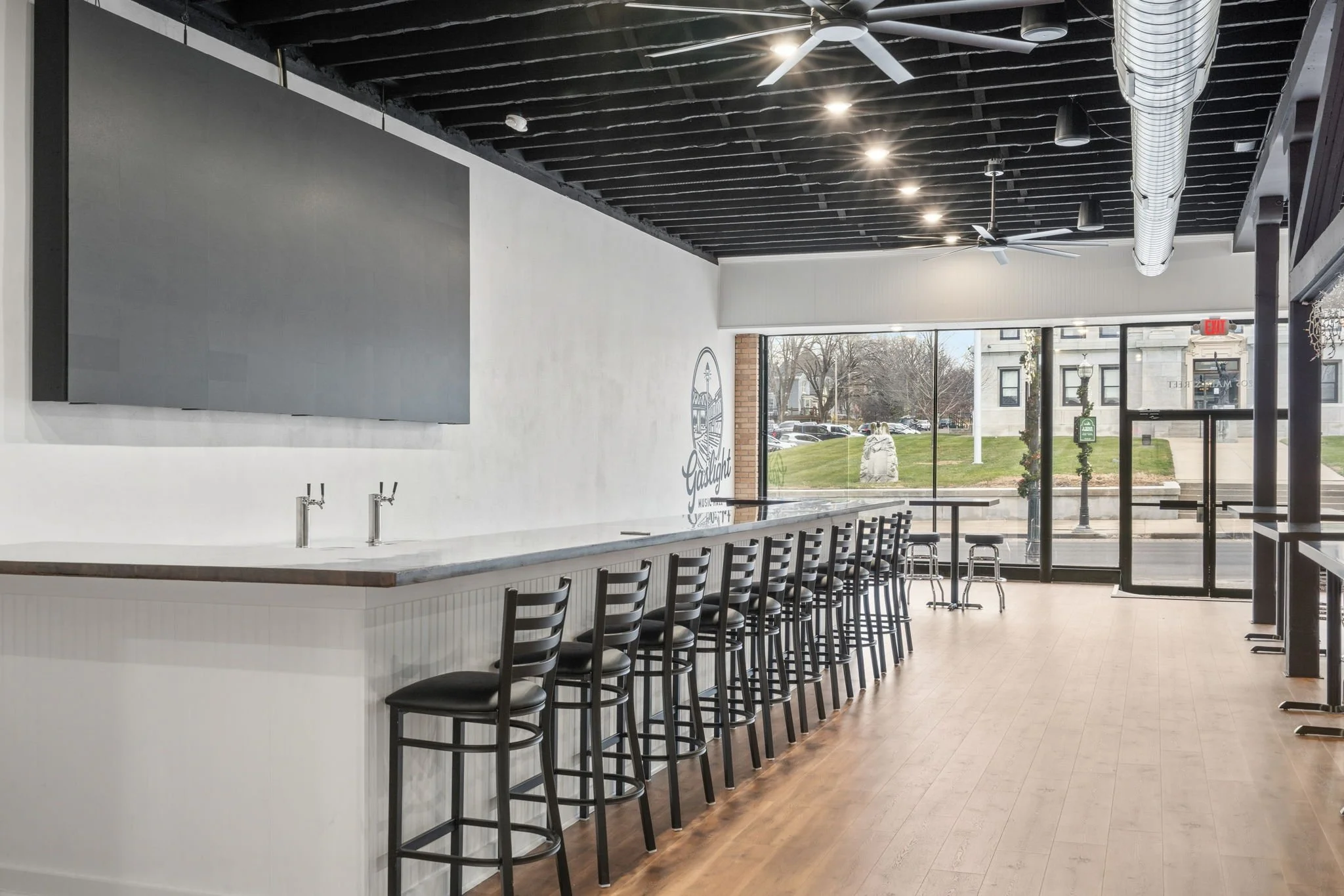 Empty modern cafe with a long white counter and black chairs, large windows, ceiling fans, and outdoor view.
