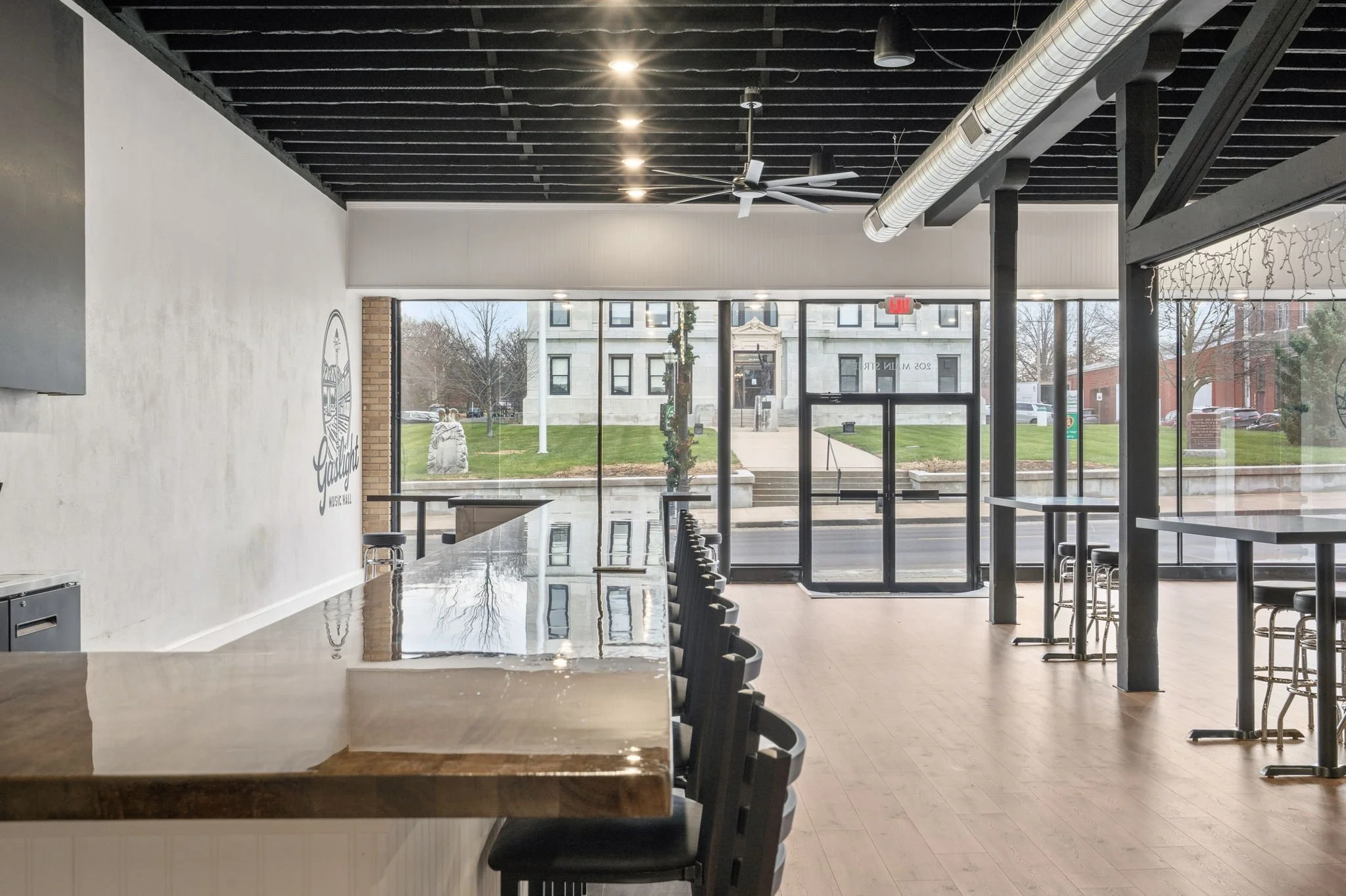 Empty interior of a modern cafe or bar with high tables and chairs, large glass front windows, and a view of a street and grassy area outside.