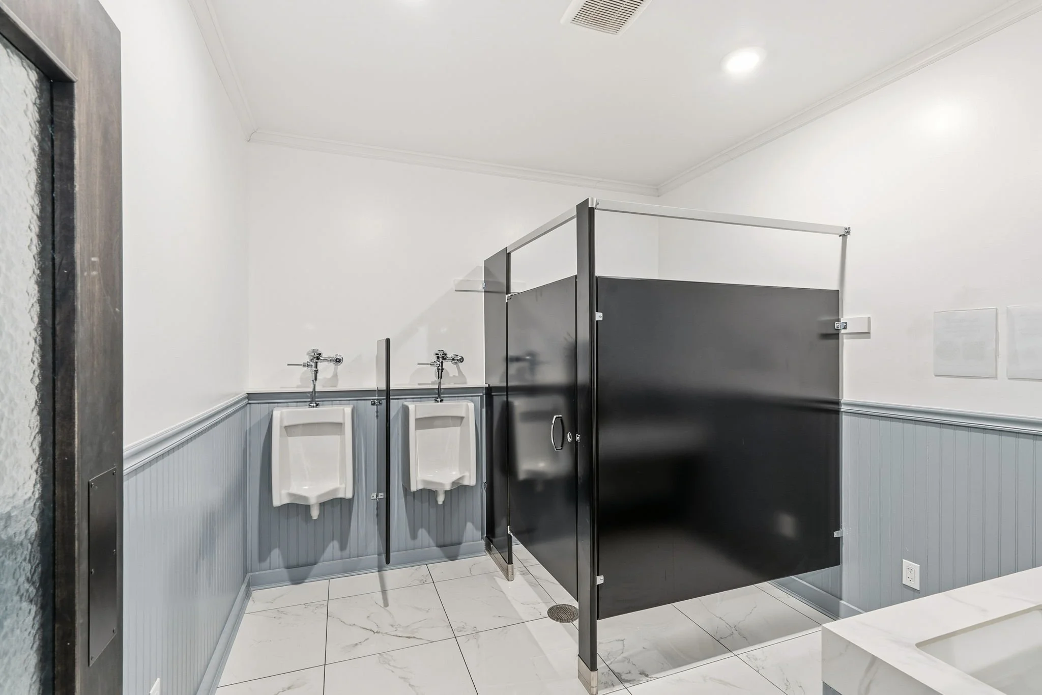 Men's bathroom with two urinals, a black privacy stall, and white tiled flooring.