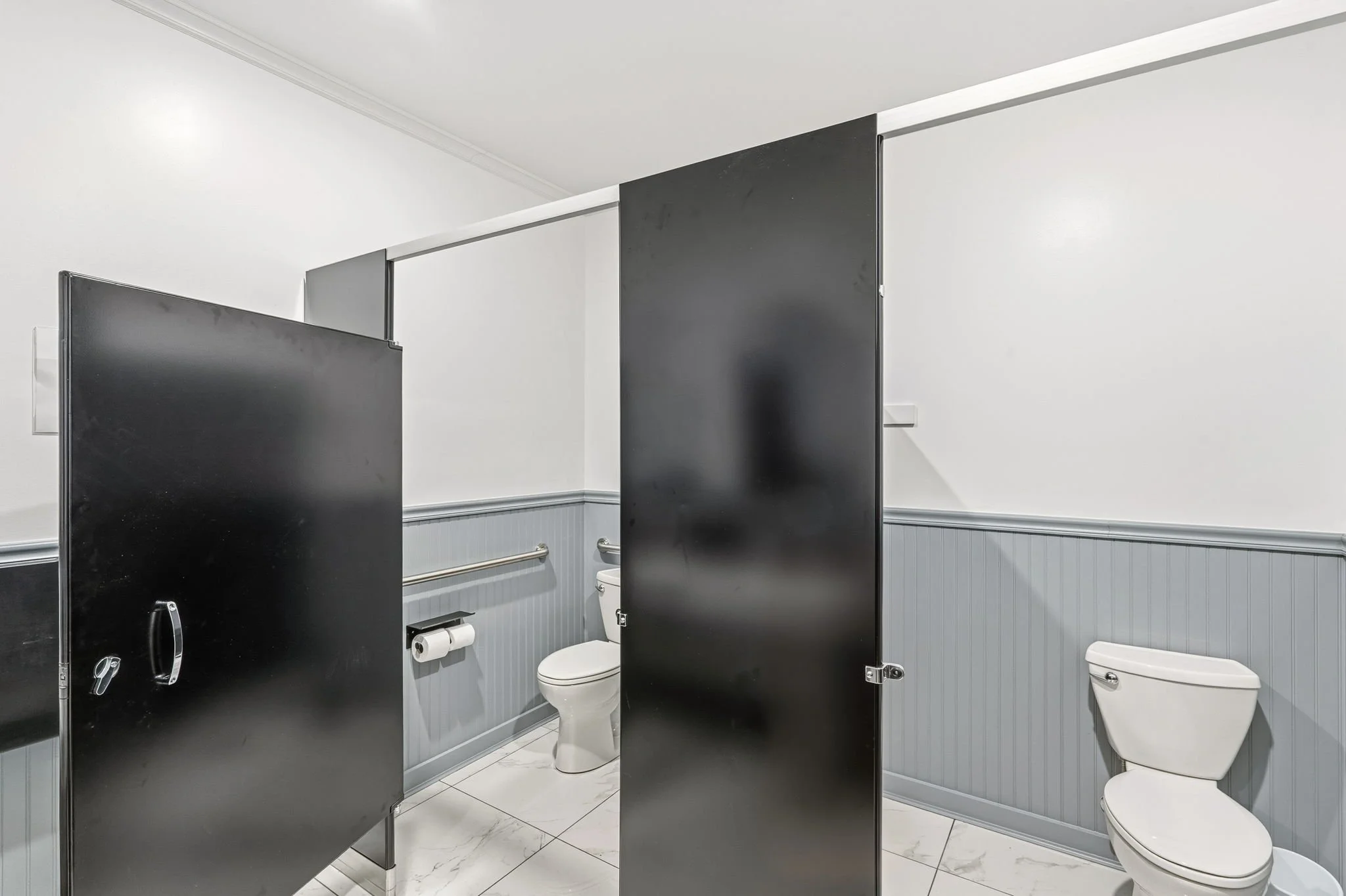 Public restroom with two toilet stalls, black stall doors, white walls, and gray wainscoting.