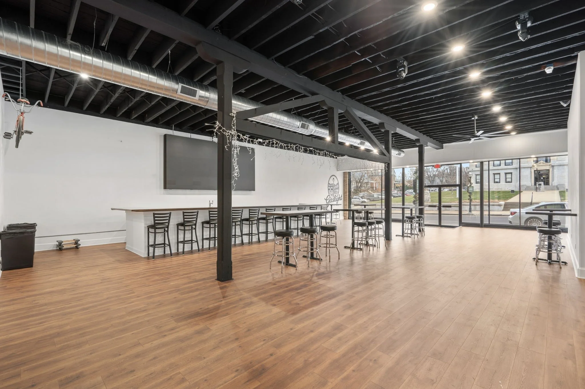 Empty modern cafe or restaurant interior with wood flooring, bar seating, high tables with stools, large windows, and ceiling lights.