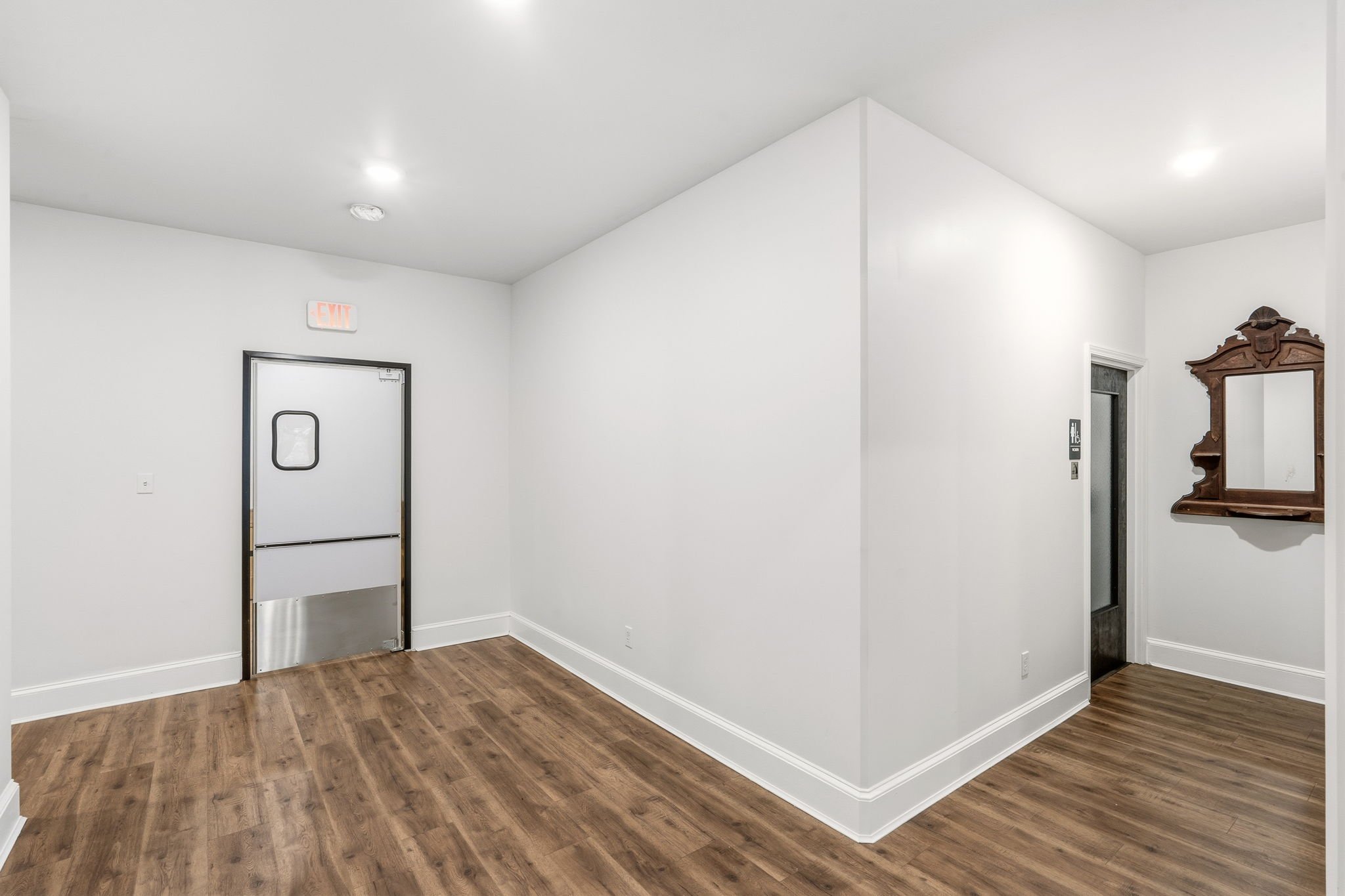 Empty room with white walls, wooden floor, metal exit door with window, and elevator door with mirror on the right wall.