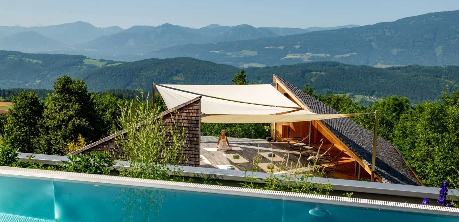 Frau meditiert auf Yogaplattform, im Vordergrund ein Pool mit Aussicht ins Gebirge