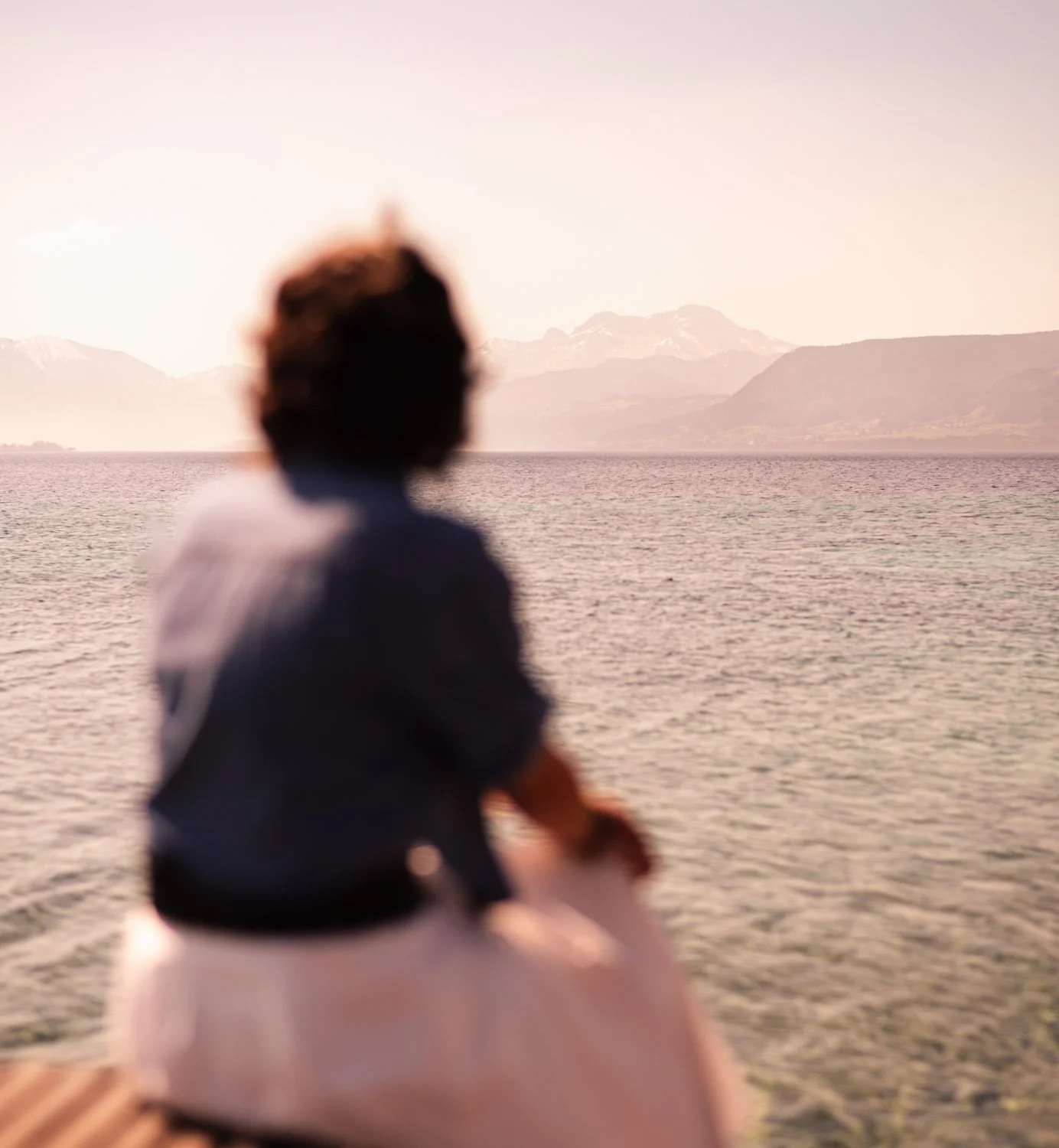 Frau sitzt am See und blickt zum Horizont, der aus einer Gebirgskette besteht
