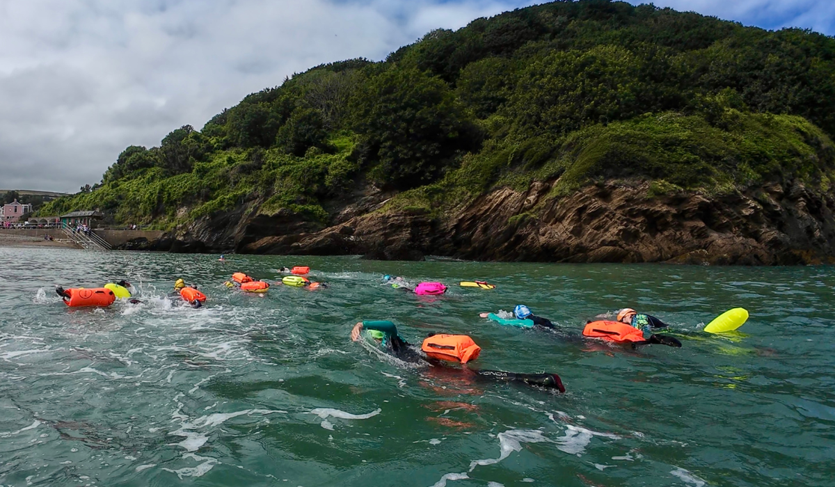 Group of people swimming in the ocean near a green hillside, wearing life jackets and swim caps.