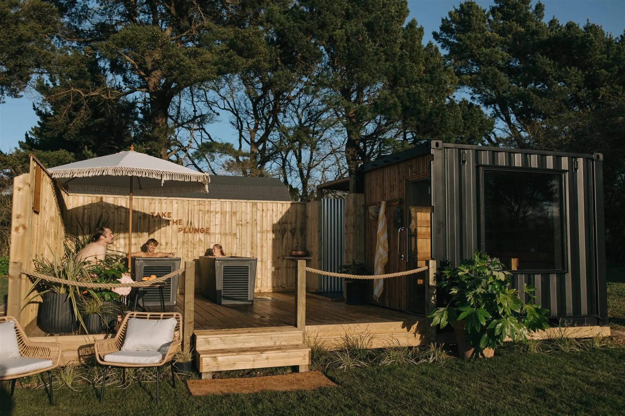 People relaxing outdoors under a white umbrella on a wooden deck with chairs and plants, surrounded by a fence and trees.