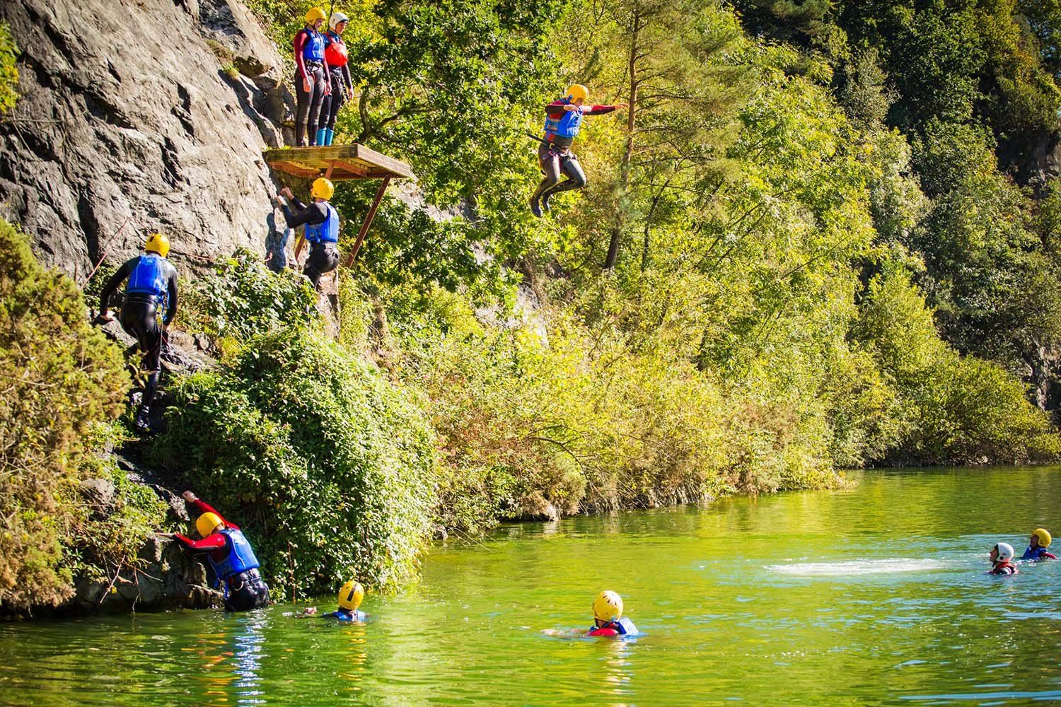 People in safety gear participating in a canyoning adventure, climbing a rocky cliff and jumping into a river surrounded by green trees.