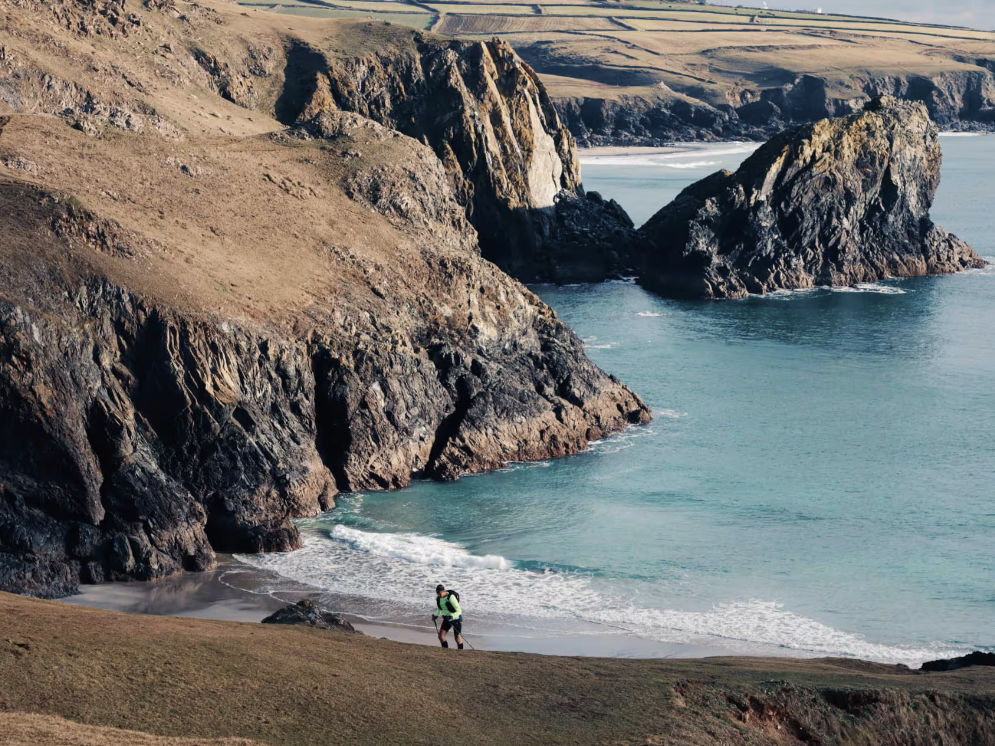 A person hiking near the shoreline at a rugged coastline with large cliffs and the ocean in the background.