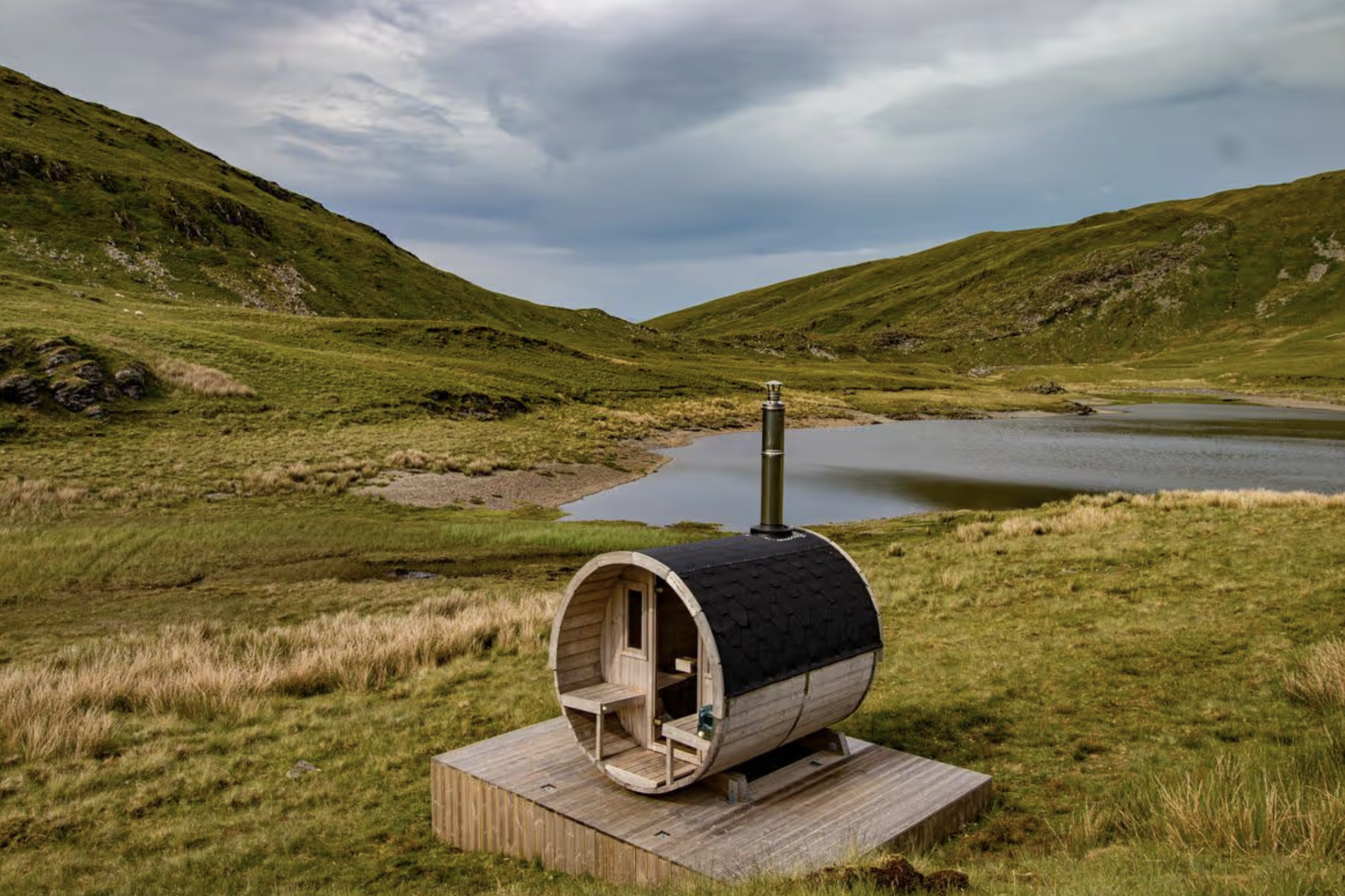 A small wooden sauna shaped like a barrel with a curved roof and a door, situated in a grassy field at the edge of a lake with rolling green hills in the background and cloudy sky overhead.