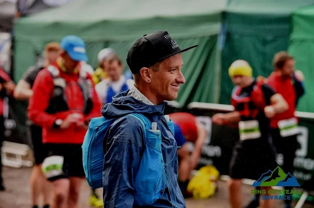 A man with a backpack and a hat stands among runners at a trail race event, with a green tent in the background.
