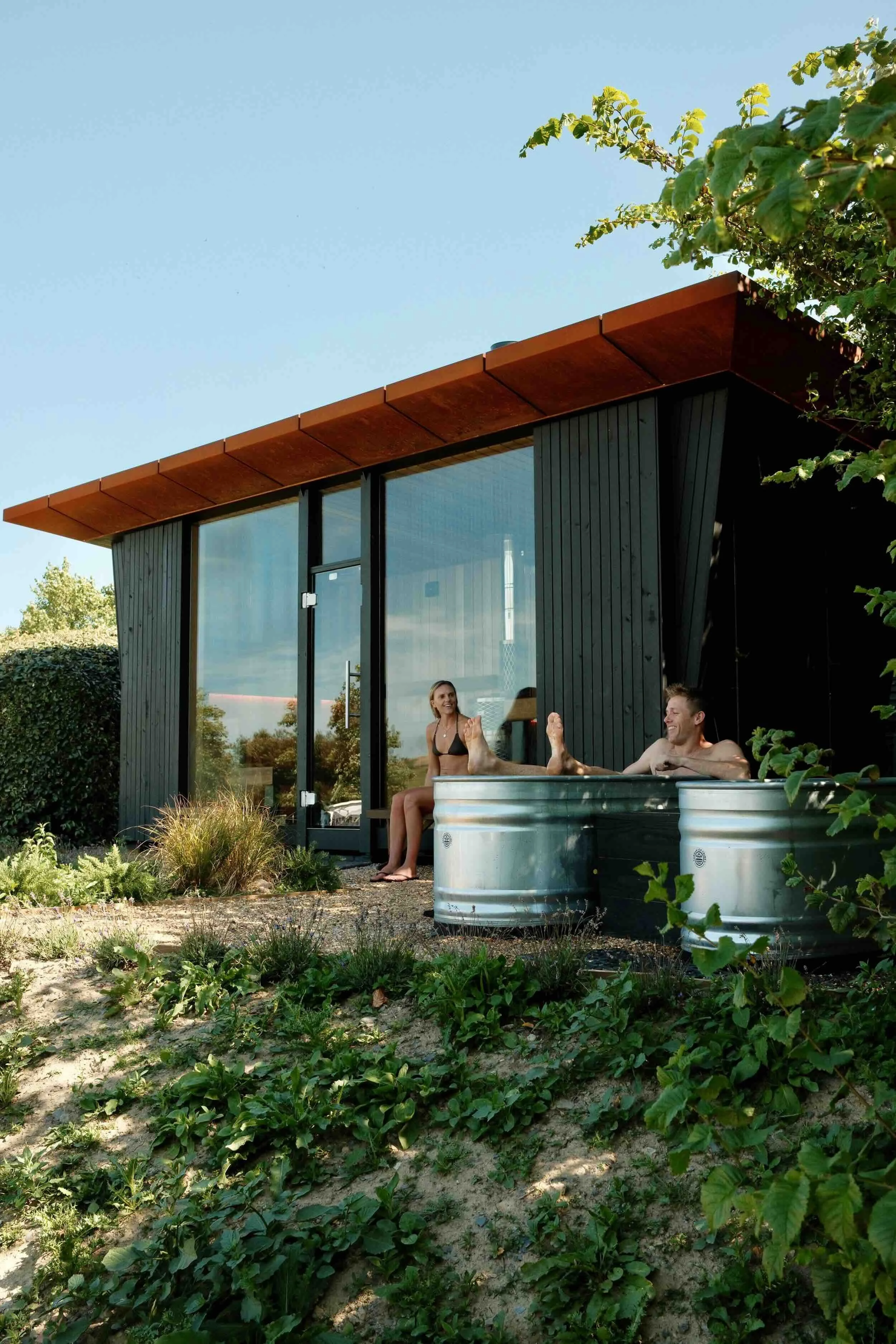 A modern house with large glass doors and a brown overhang, surrounded by greenery. Two people are relaxing in a small above-ground pool in the yard.