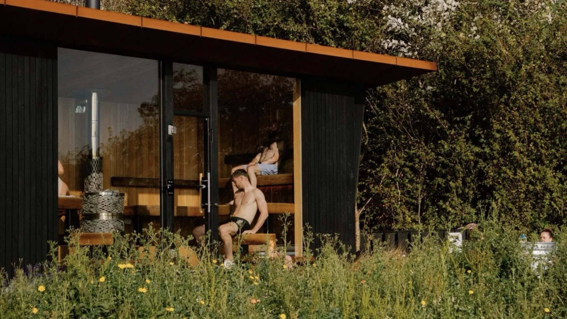 Modern sauna with glass front, located outdoors among trees and wildflowers. Inside, a shirtless man is sitting on a bench, wearing shorts, while a woman is sitting above him on the upper bench. In the outdoor area, there is a table and a barrel, with a person partially visible on the right side of the image.