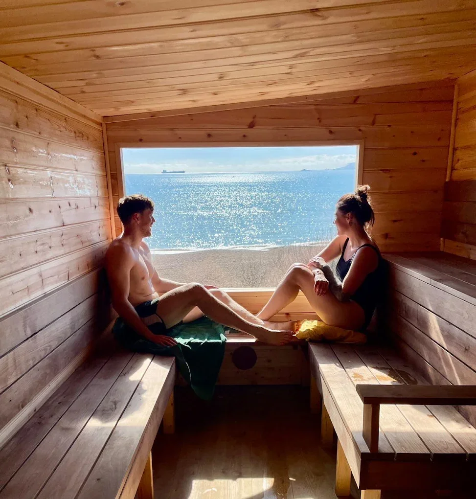 A man and woman sitting in a wooden sauna with a large window overlooking the ocean and a sandy beach.
