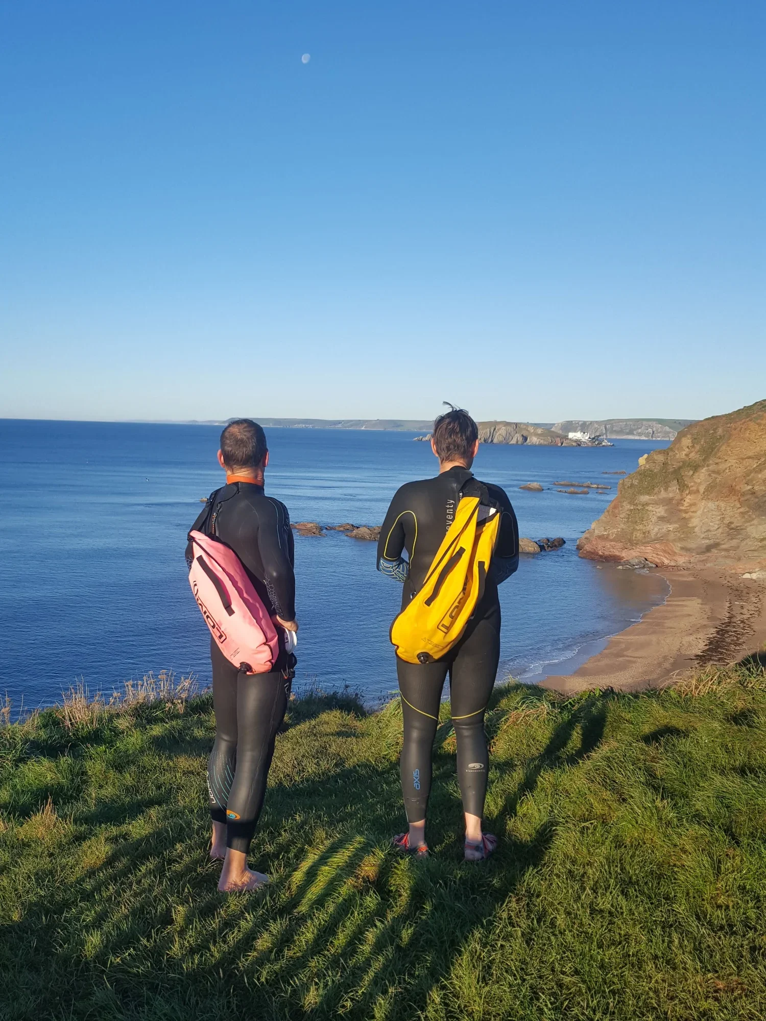 Two people in wetsuits holding wetsuit bags, standing barefoot on grass near the ocean with cliffs in the background, under a clear blue sky with the moon visible.