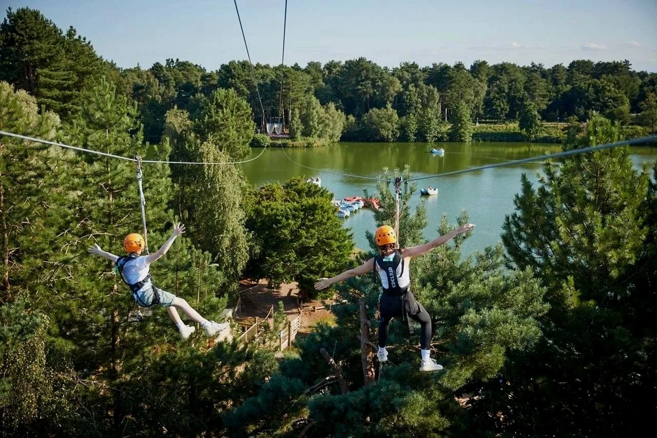 Two people wearing helmets and harnesses are zip-lining above a wooded area with a lake in the background, with boats on the water and trees surrounding the lake.