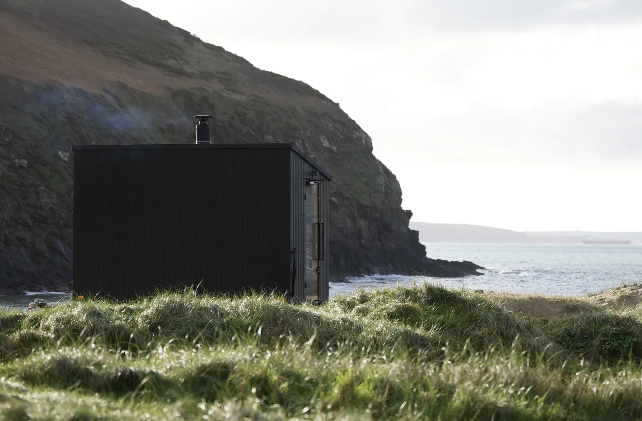 Black modern tiny house by the ocean with steep hill in background, cloudy sky, grass in foreground.