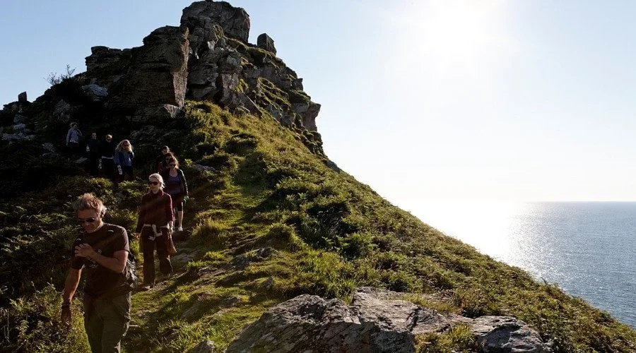 A group of people hiking along a grassy trail on a hillside near the ocean with rocky formations.
