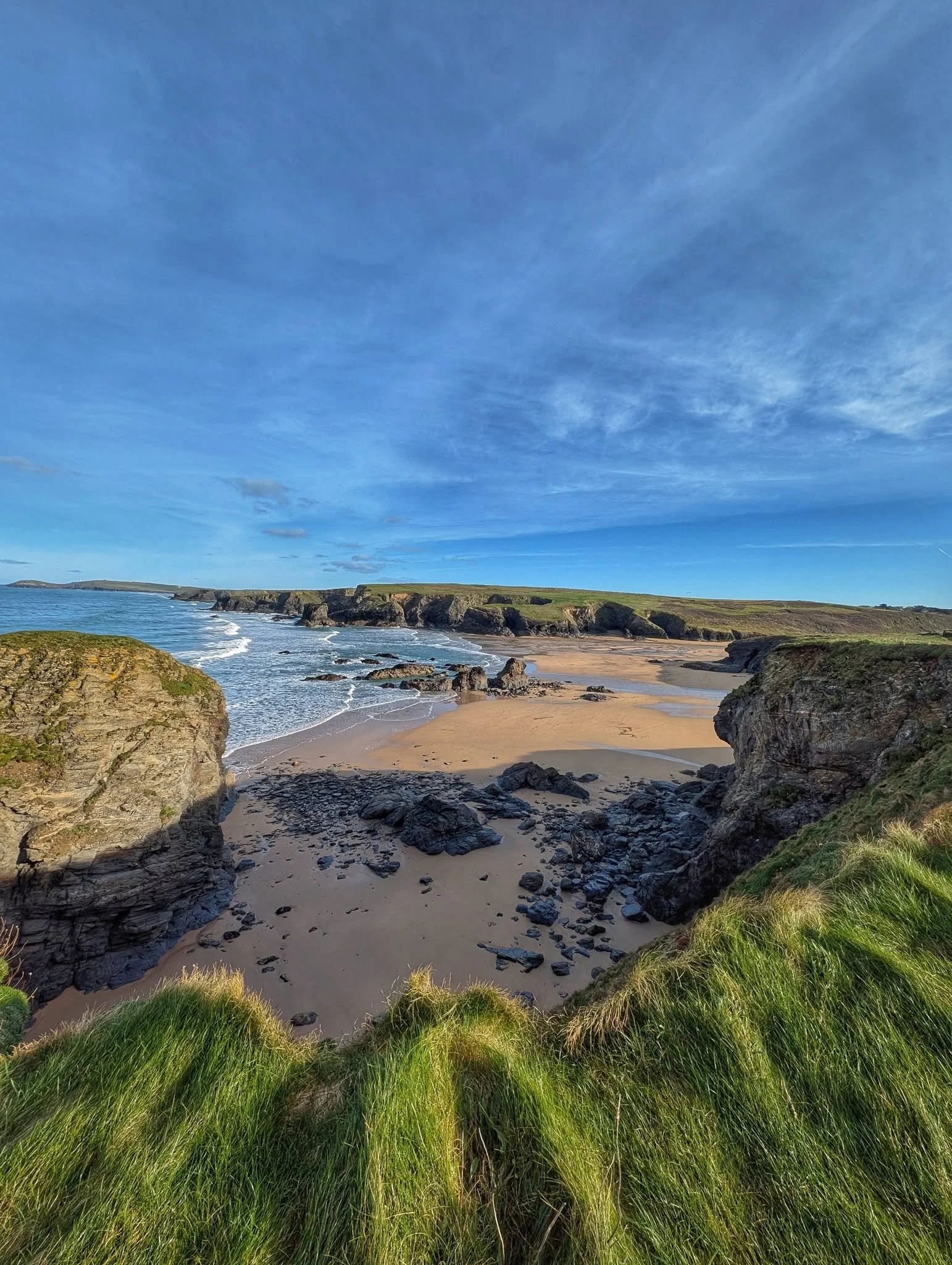 A coastal scene with grassy foreground, rocky cliffs, sandy beach, and gentle ocean waves under a blue sky with wispy clouds.
