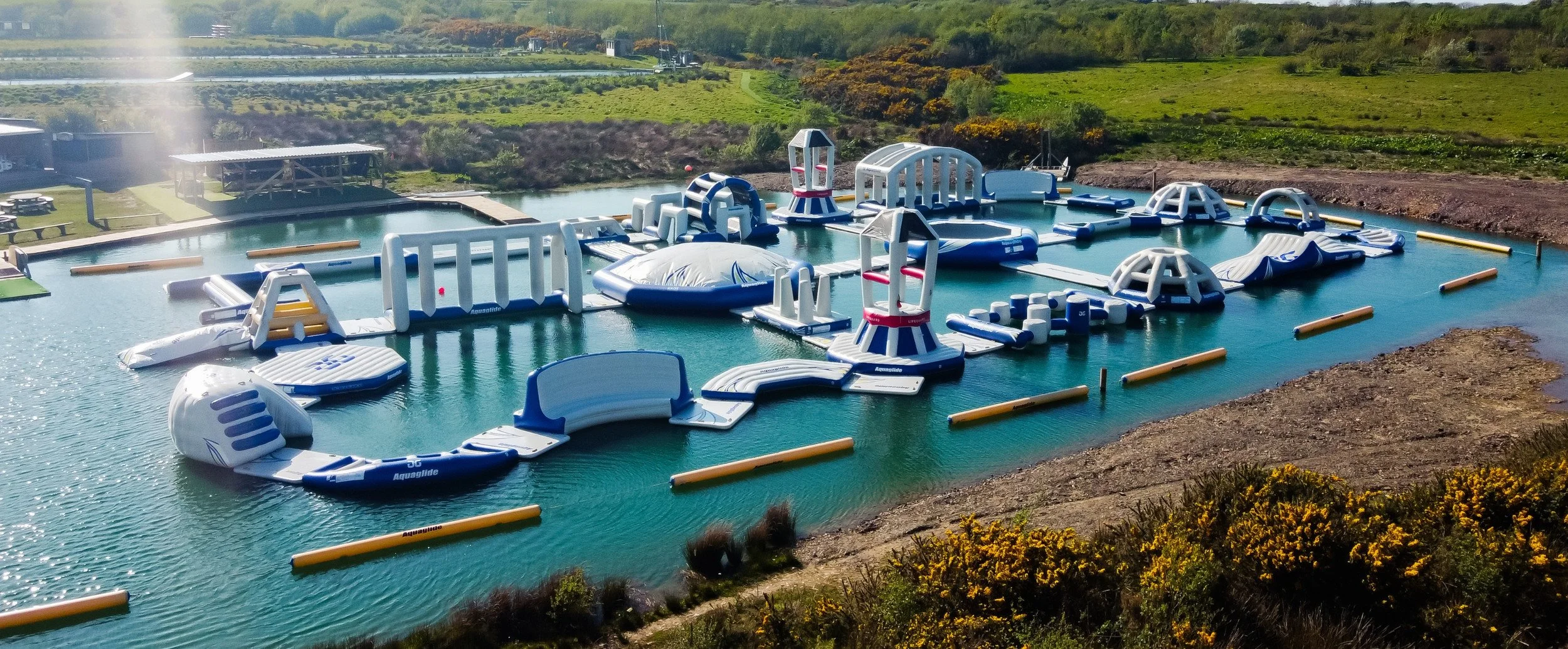 An outdoor inflatable water park with various inflatable structures and slides on a body of water, surrounded by green landscape and some shrubs in the foreground.