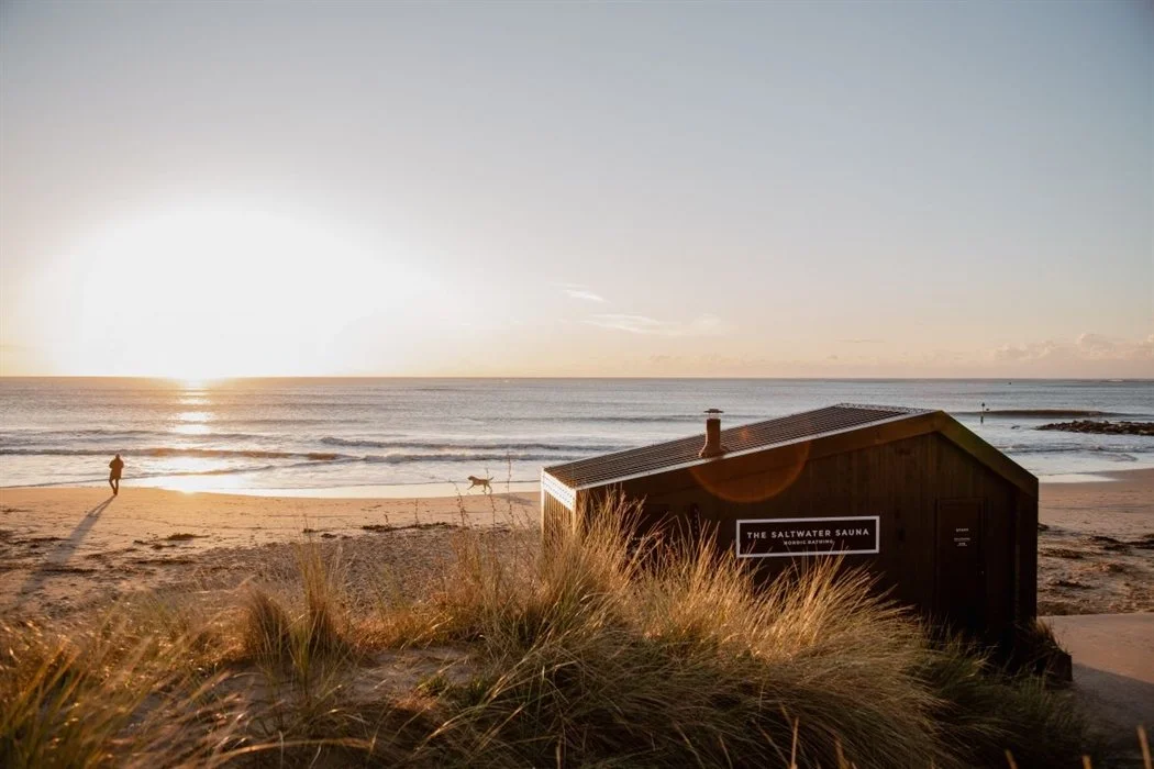 Sunset over the ocean with a beach in the foreground, a person walking a dog, and a black sauna building labeled "The Saltwater Sauna" near sand dunes and tall grass.
