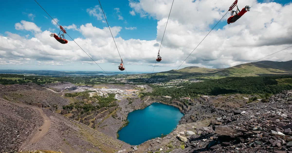 Four harnessed workers on zip lines are suspended above a rocky landscape with a small lake, industrial area, and green hills in the background under a partly cloudy sky.