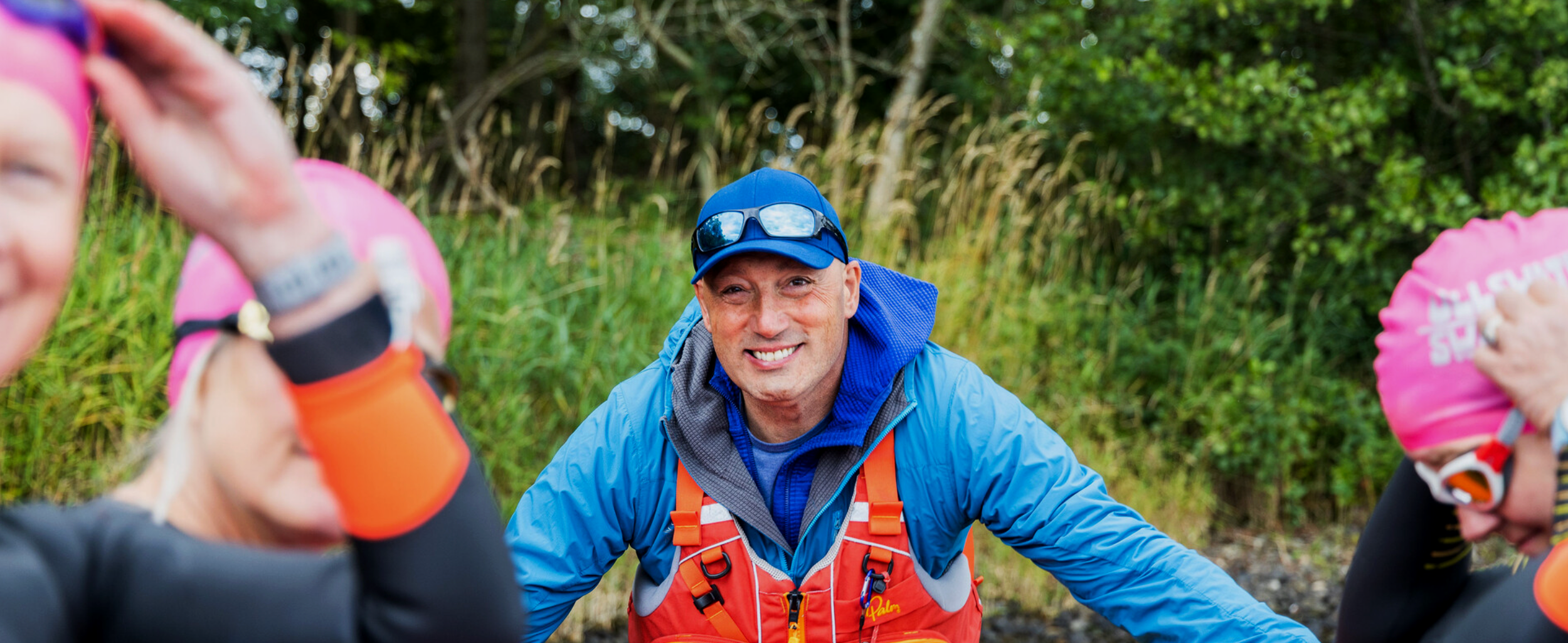 Smiling man in blue jacket and cap talking with two women in pink hats outdoors with green foliage.