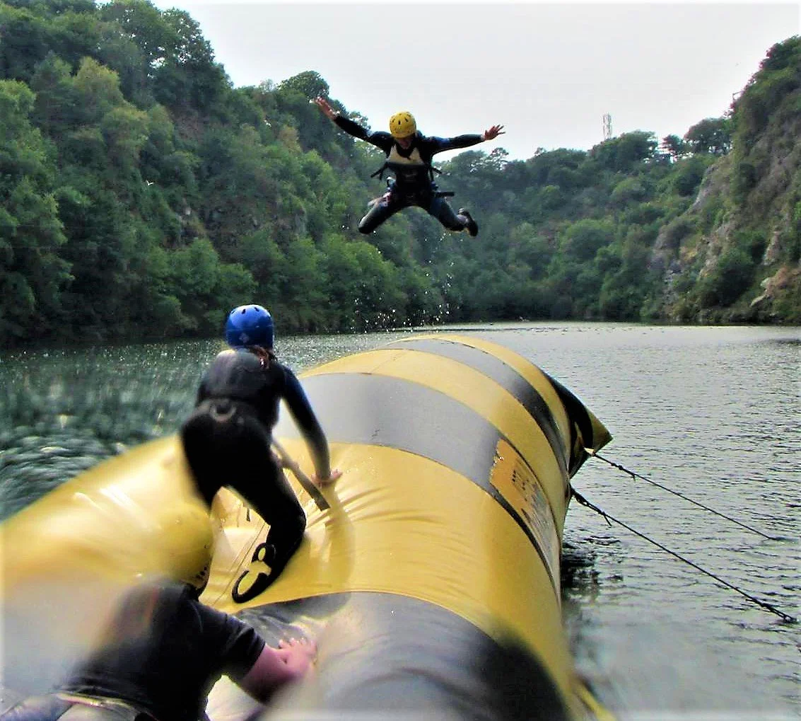 Two people on a yellow submerged pipe in water, with one person climbing, and the other jumping into the water wearing helmets. Lush green trees on both sides of the waterway.