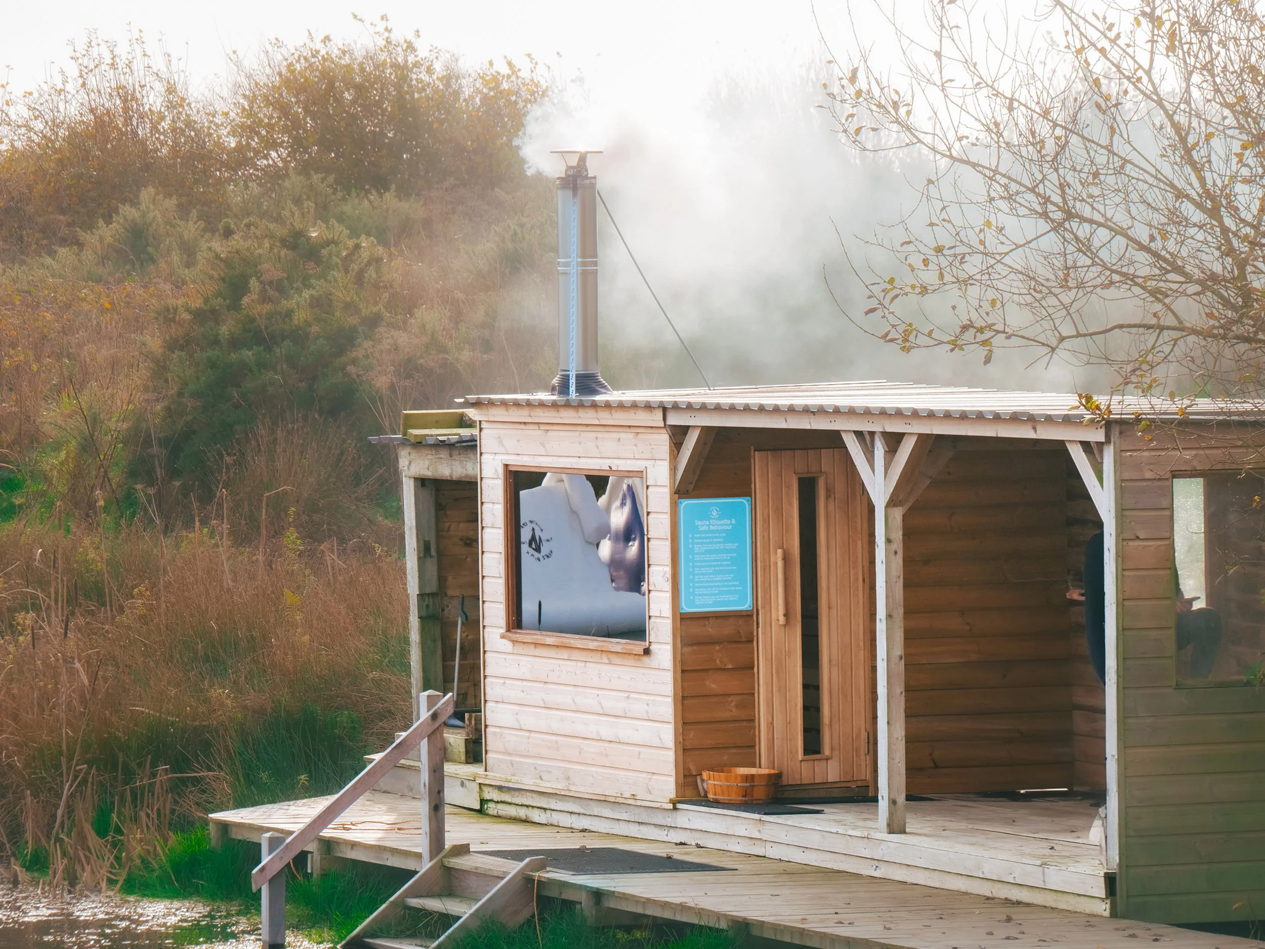 Wooden sauna shed with smoke rising from chimney, surrounded by autumn trees and grass, on a wooden deck with stairs.