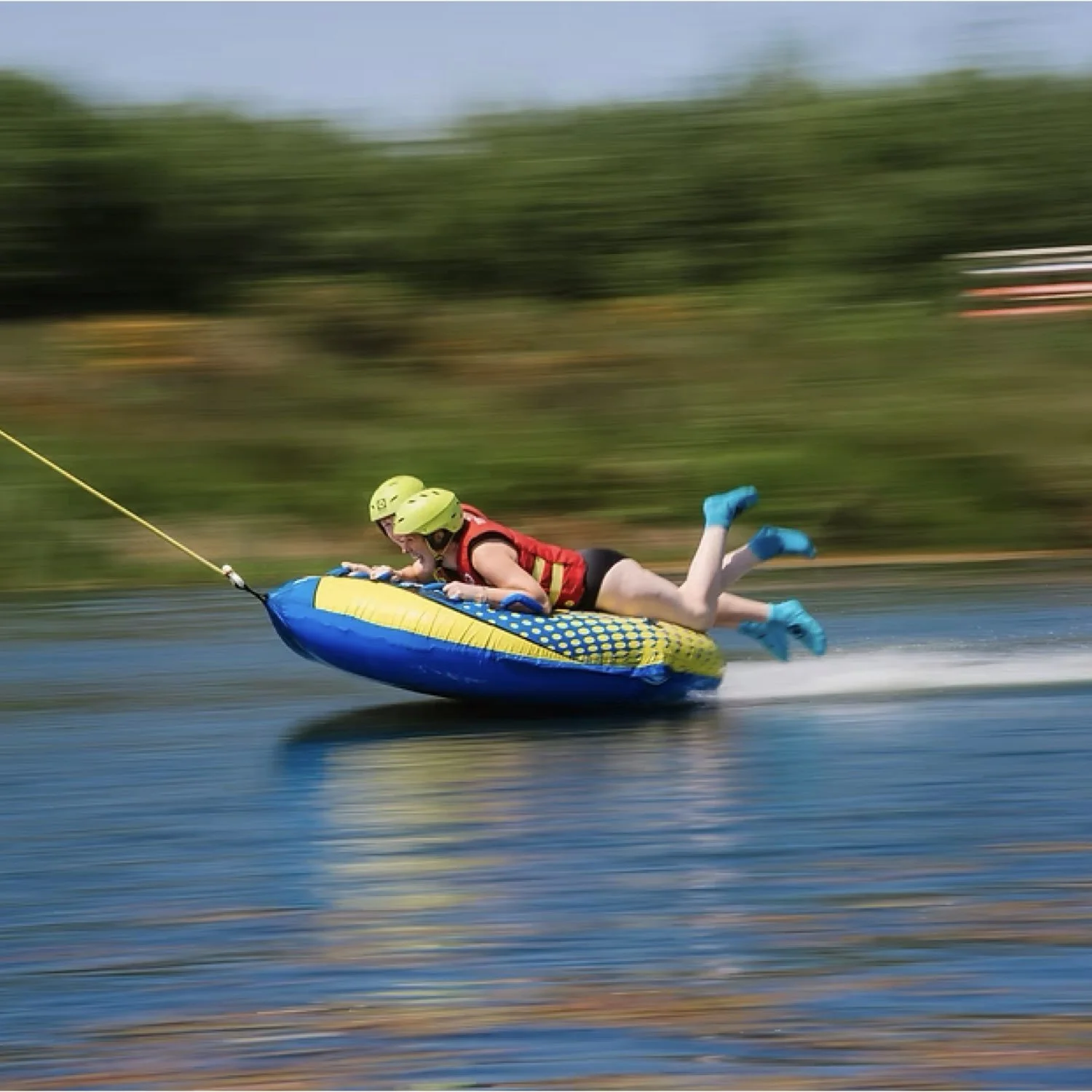 Child in a red life jacket and green helmet riding an inflatable water tube, being pulled by a boat on a lake, with trees in the background.