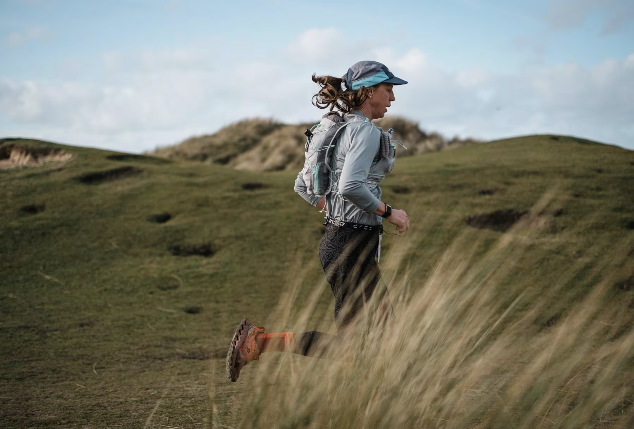 A woman trail running on a grassy hillside, wearing a gray jacket, black patterned leggings, a blue and gray cap, and a hydration pack.