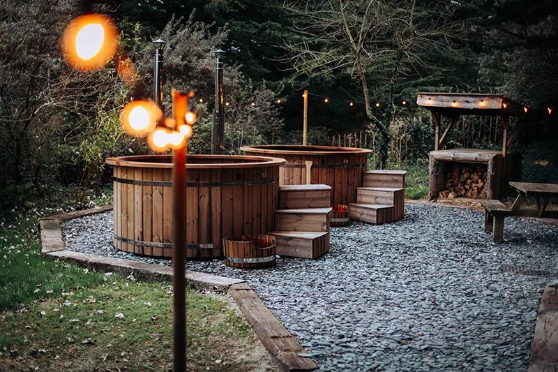 Outdoor hot tubs with wooden steps in a backyard, surrounded by trees and string lights at dusk.