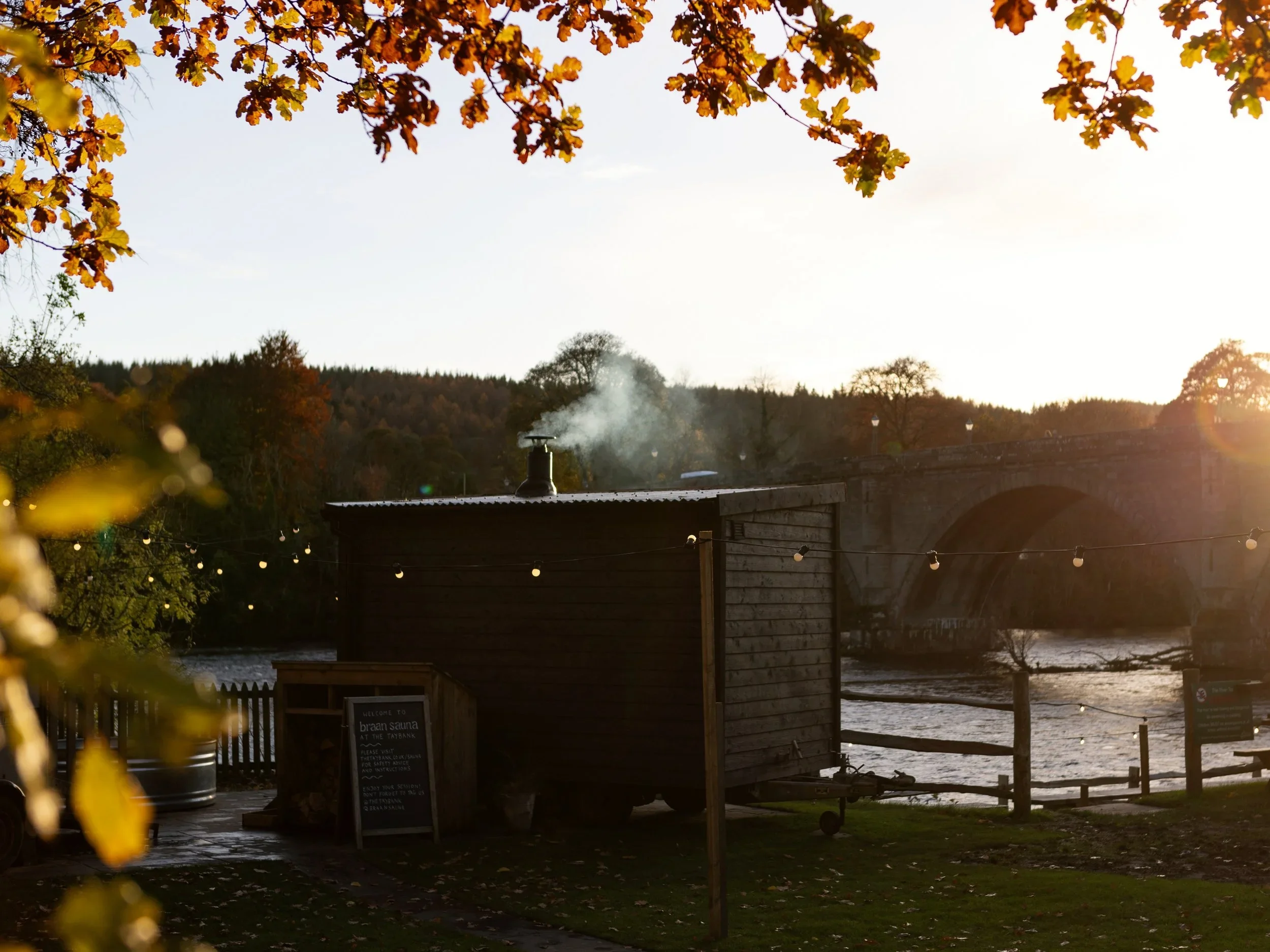 A riverside scene during sunset with trees displaying autumn foliage. In the foreground, there is a small building with a chimney emitting smoke and a sign for a sauna. String lights are hanging, and a stone bridge crosses the river in the background.
