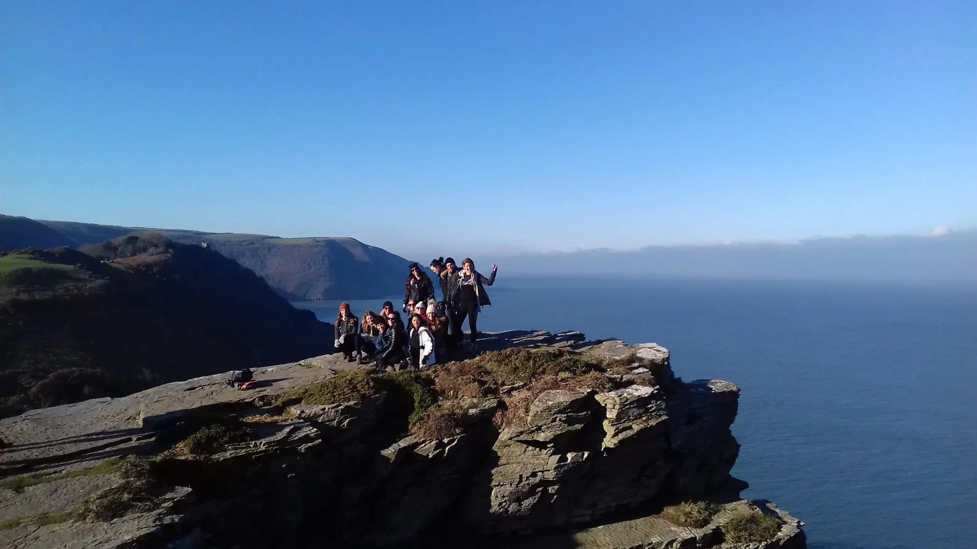 Group of people hiking and posing on a rocky cliff overlooking the ocean with cliffs in the background under a clear blue sky.