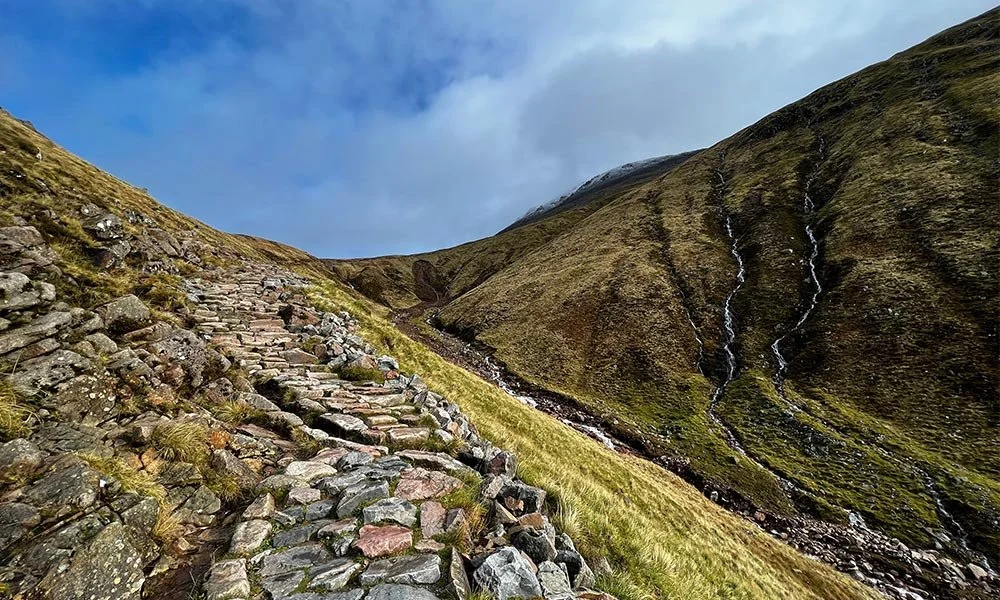 A rugged stone pathway winds up a grassy hillside with mountains and a cloudy sky in the background.