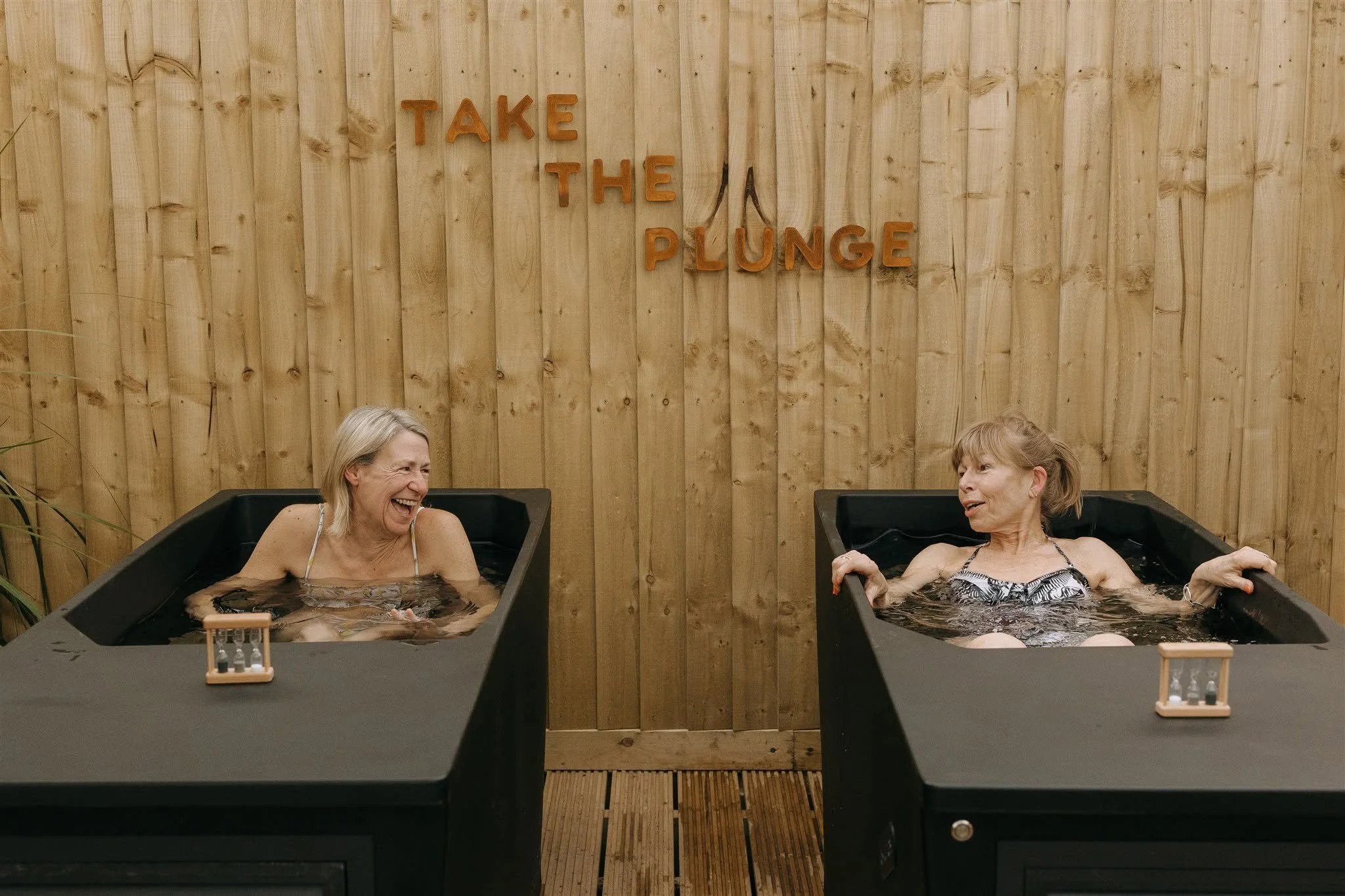 Two women in hot tubs, laughing and talking, with a wooden wall background and a sign that says "Take the Plunge."