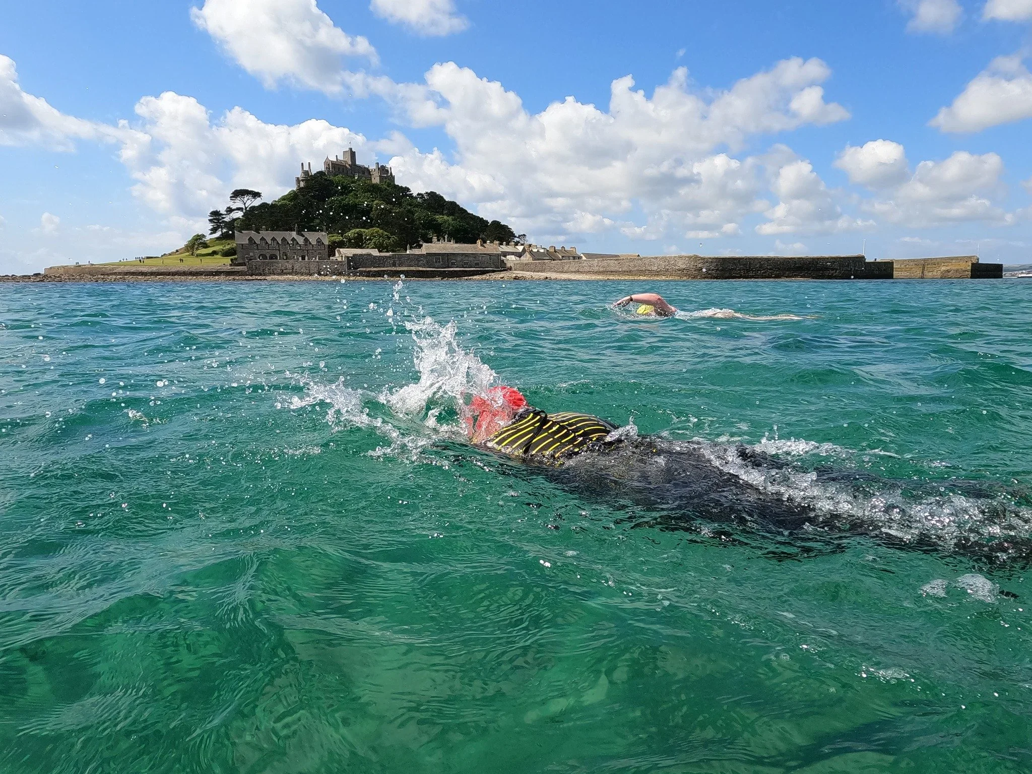 A swimmer in a black and yellow swimsuit swimming in the ocean near a rocky island with buildings and trees on top, under a partly cloudy sky.