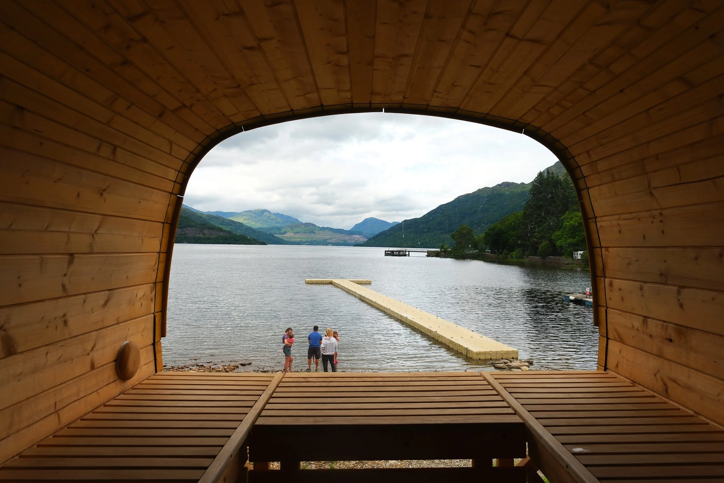 View of a lake and mountains through a wooden arch structure with a curved opening and bench inside, with four people standing near the water.