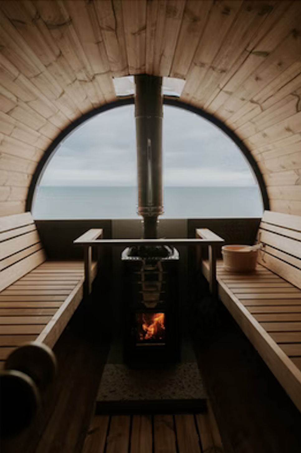 Interior of a small wooden sauna with bench seating, a pot on the right bench, a stove with fire in the center, and a large arched window showing an ocean view.