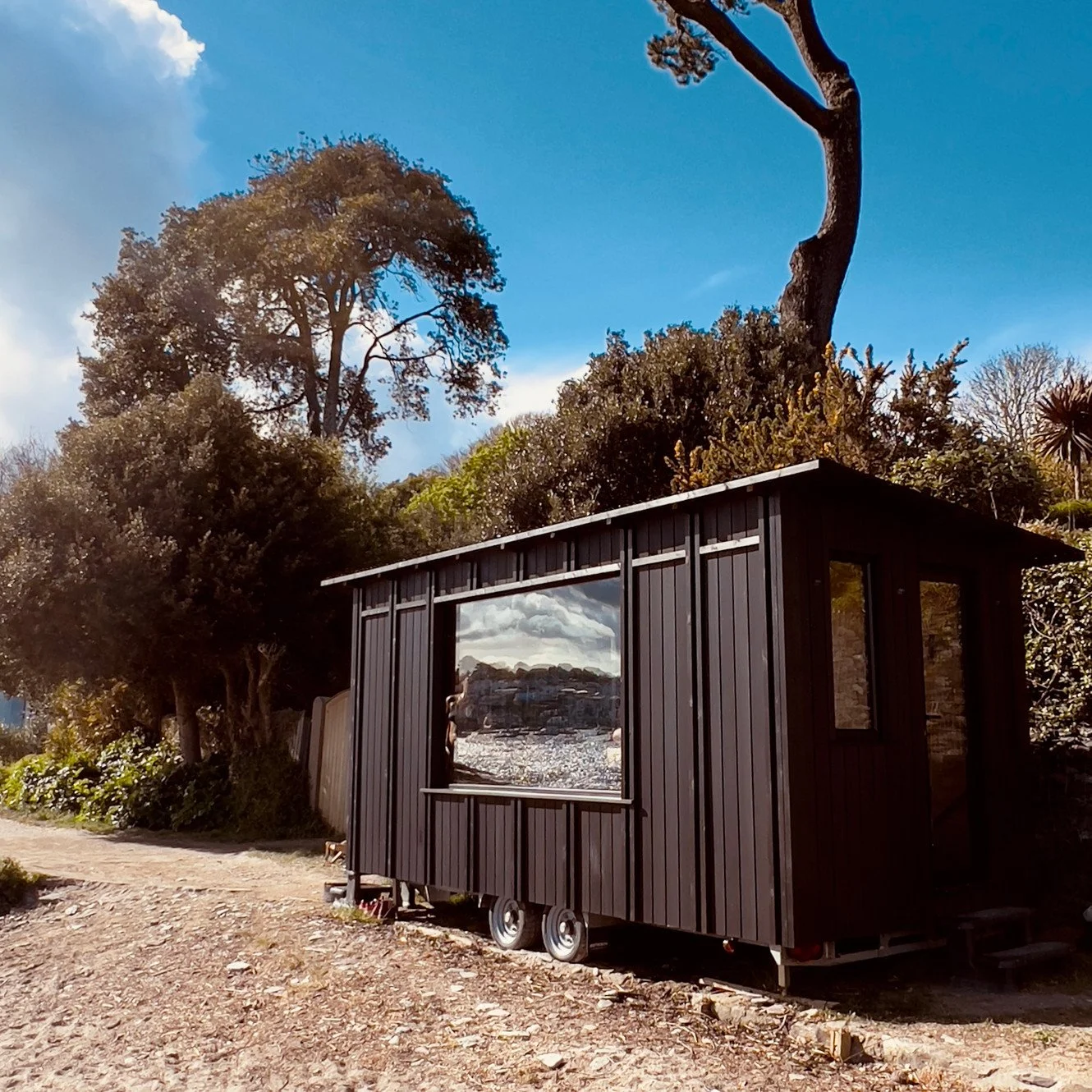 A small black tiny house on wheels with a large side window reflecting clouds and mountains, situated on a dirt path surrounded by trees and bushes under a blue sky.