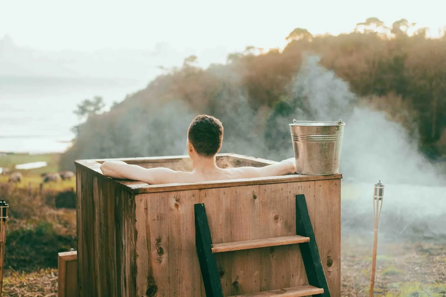 A person relaxing in a wooden hot tub outdoors, overlooking a scenic natural landscape with trees and a river, during sunrise or sunset.