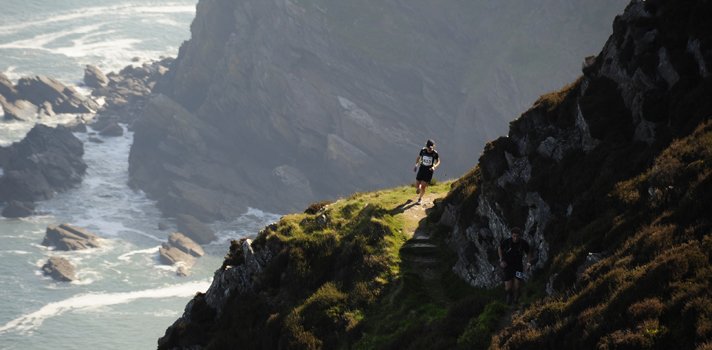 Two people hiking on a narrow mountain trail along a steep cliffside overlooking the ocean with waves crashing below.