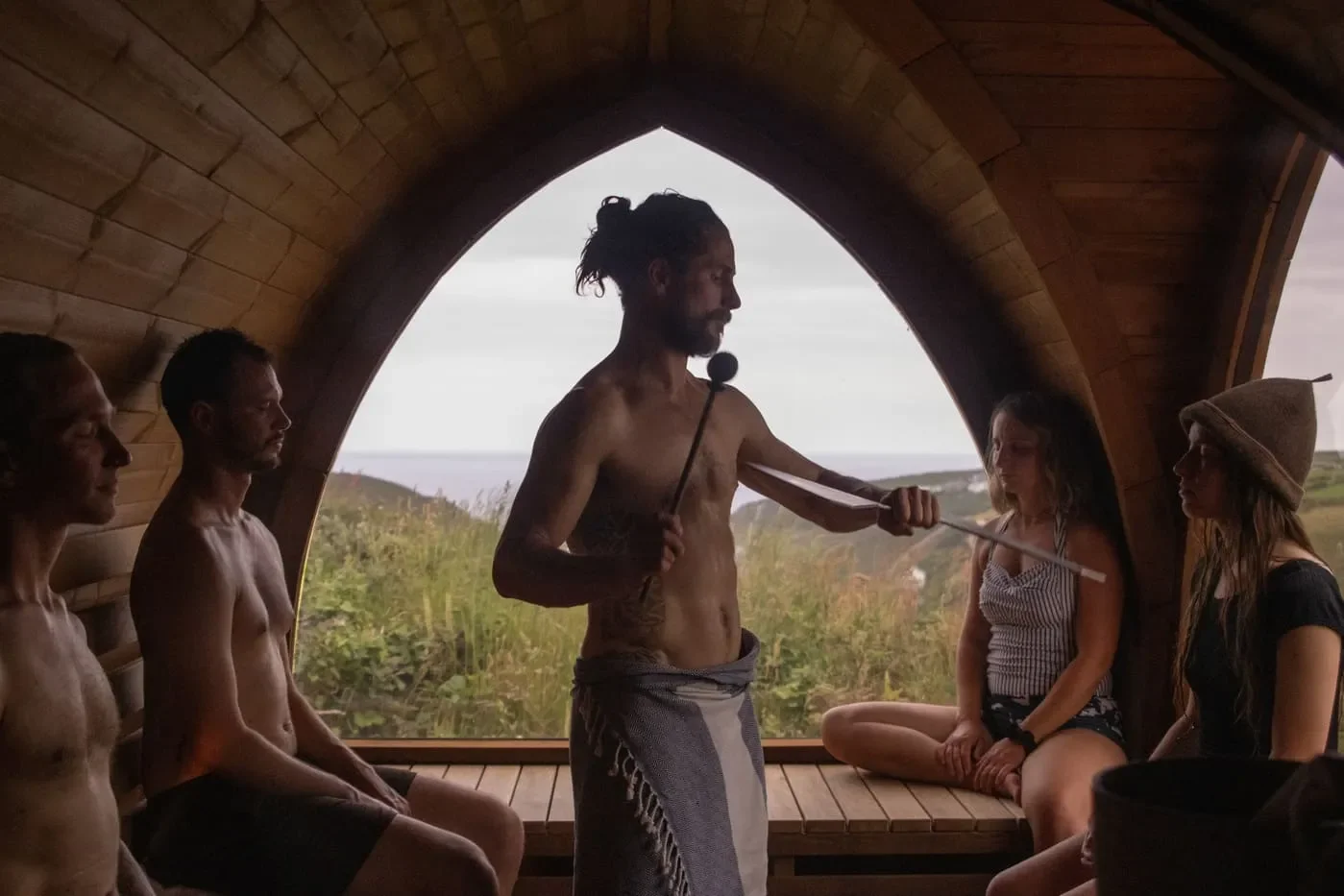 A man with long, curly hair and a beard, shirtless, leading a meditation or sound therapy session inside a wooden dome with five seated participants, three men and two women, overlooking a natural landscape.